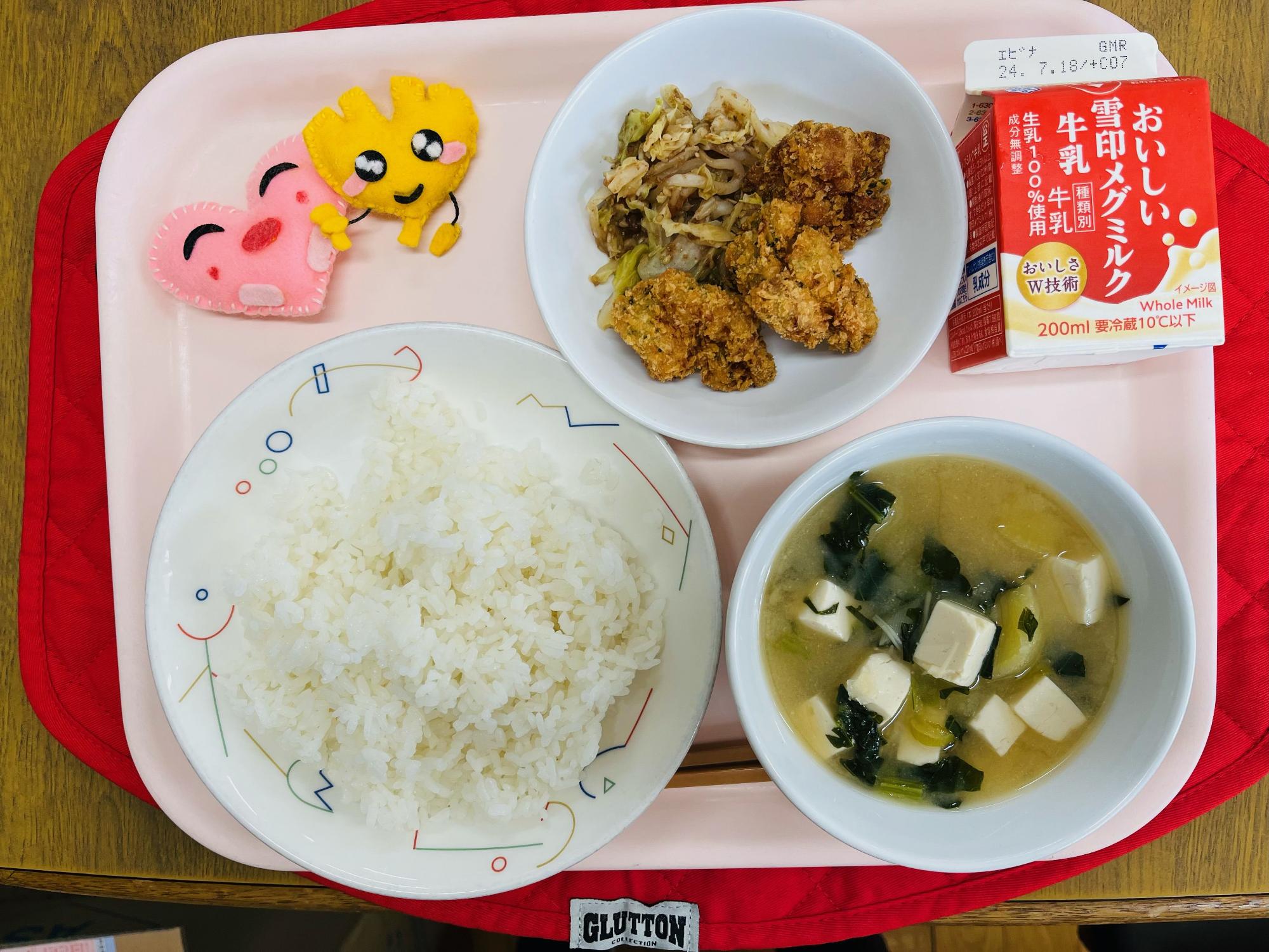 ピンクのトレーに給食が右上から反時計回りで牛乳、きゃべつのおかかあえとチキンカツ、ハートの形をした黄色とピンク色の布製マスコット、ごはん、味噌汁と並べられている写真