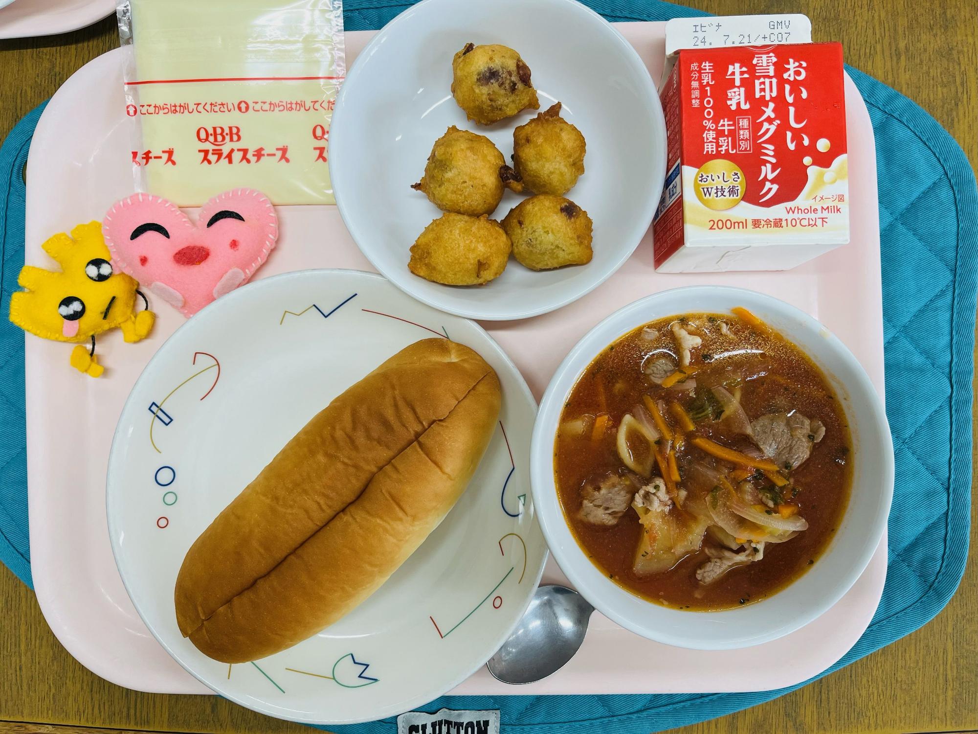 ピンクのトレーに給食が右上から反時計回りで牛乳、フルーツボール、チーズ、ハートの形をした黄色とピンク色の布製マスコット、サンドパン、ミネストローネと並べられている写真
