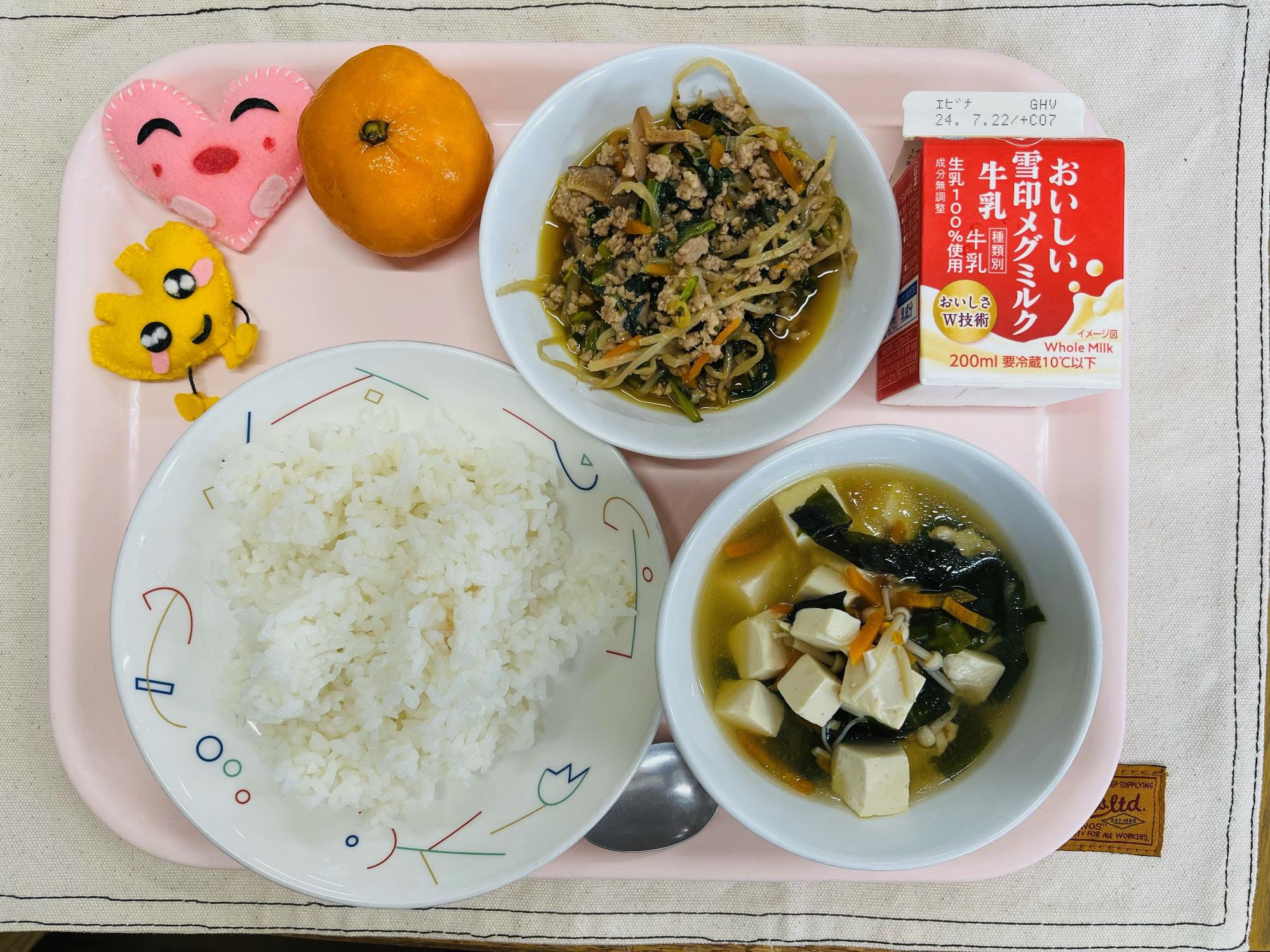 ピンクのトレーに給食が右上から反時計回りで牛乳、ビビンバ、れいとうみかん、ハートの形をした黄色とピンク色の布製マスコット、ごはん、わかめスープと並べられている写真