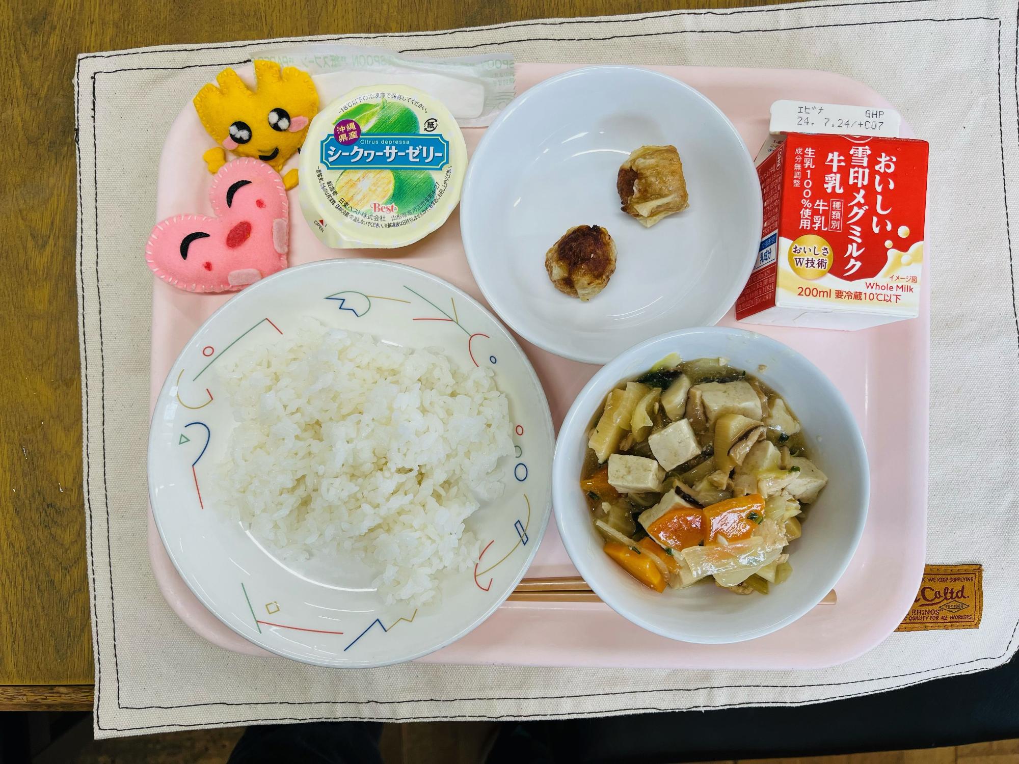 ピンクのトレーに給食が右上から反時計回りで牛乳、揚げシューマイ、シークァーサーゼリー、ハートの形をした黄色とピンク色の布製マスコット、ごはん、豆腐の中華煮と並べられている写真