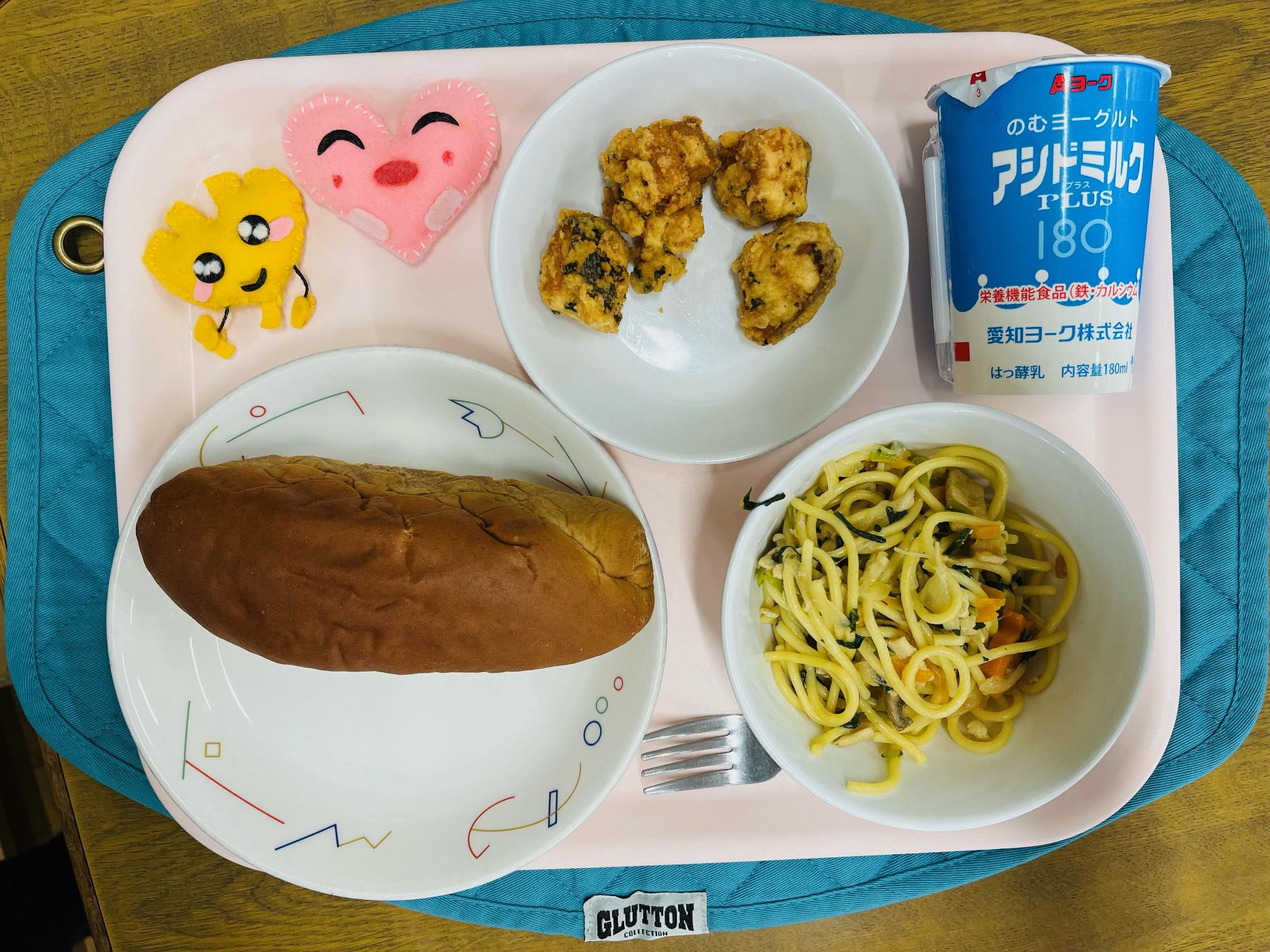 ピンクのトレーに給食が右上から反時計回りで発酵乳、さけのチーズ揚げ、ハートの形をした黄色とピンク色の布製マスコット、くろパン、和風スパゲッティと並べられている写真
