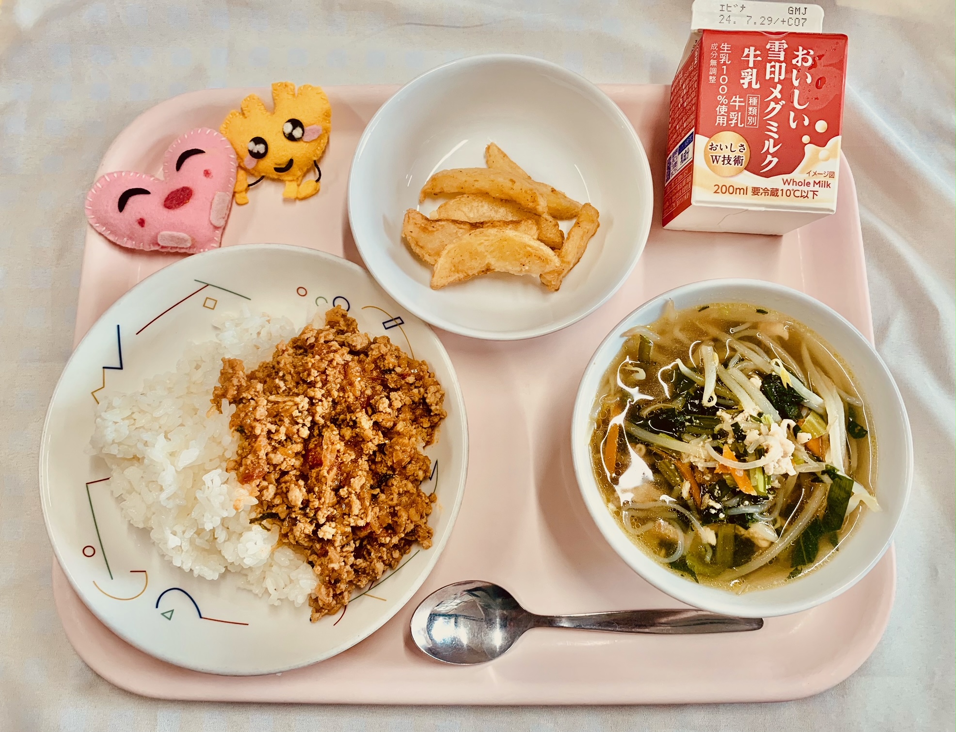 ピンクのトレーに給食が右上から反時計回りで牛乳、チリポテト、ハートの形をした黄色とピンク色の布製マスコット、トマトとひき肉のアジアンライス、五目スープと並べられている写真