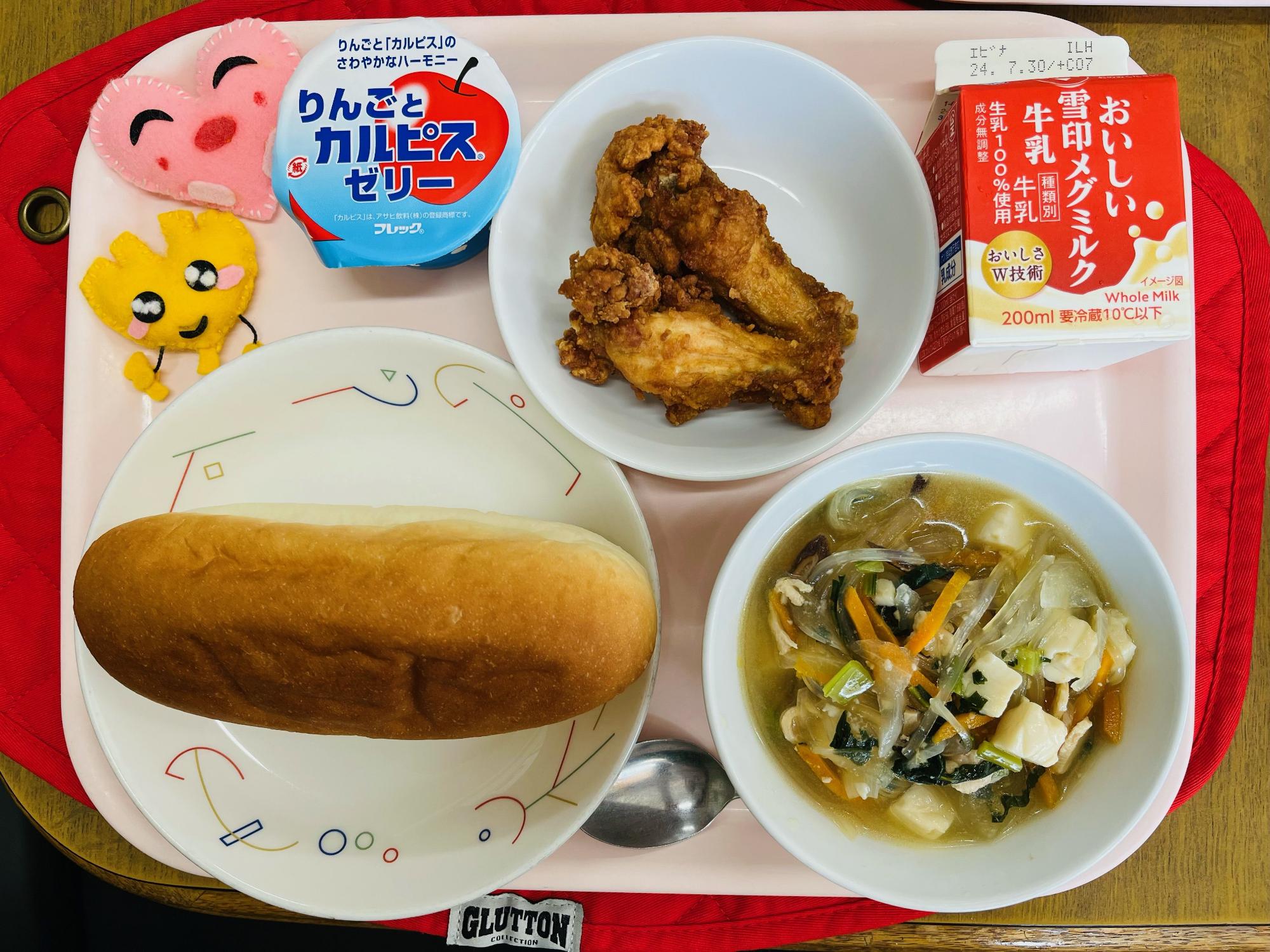 ピンクのトレーに給食が右上から反時計回りで牛乳、フライドチキン、りんごとカルピスゼリー、ハートの形をした黄色とピンク色の布製マスコット、コッペパン、春雨スープと並べられている写真