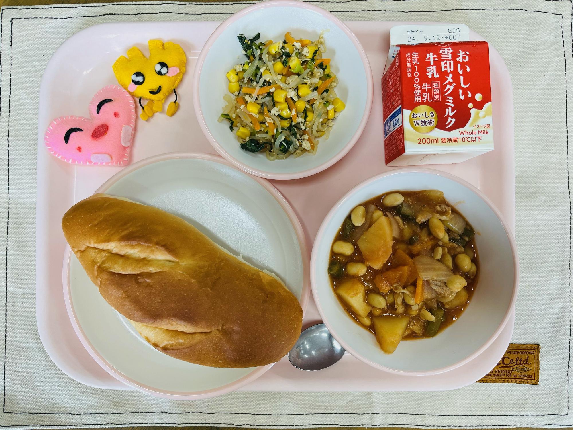 ピンクのトレーに給食が右上から反時計回りで牛乳、しらたきソテー、ハートの形をした黄色とピンク色の布製マスコット、ミルクパン、ポークビーンズと並べられている写真