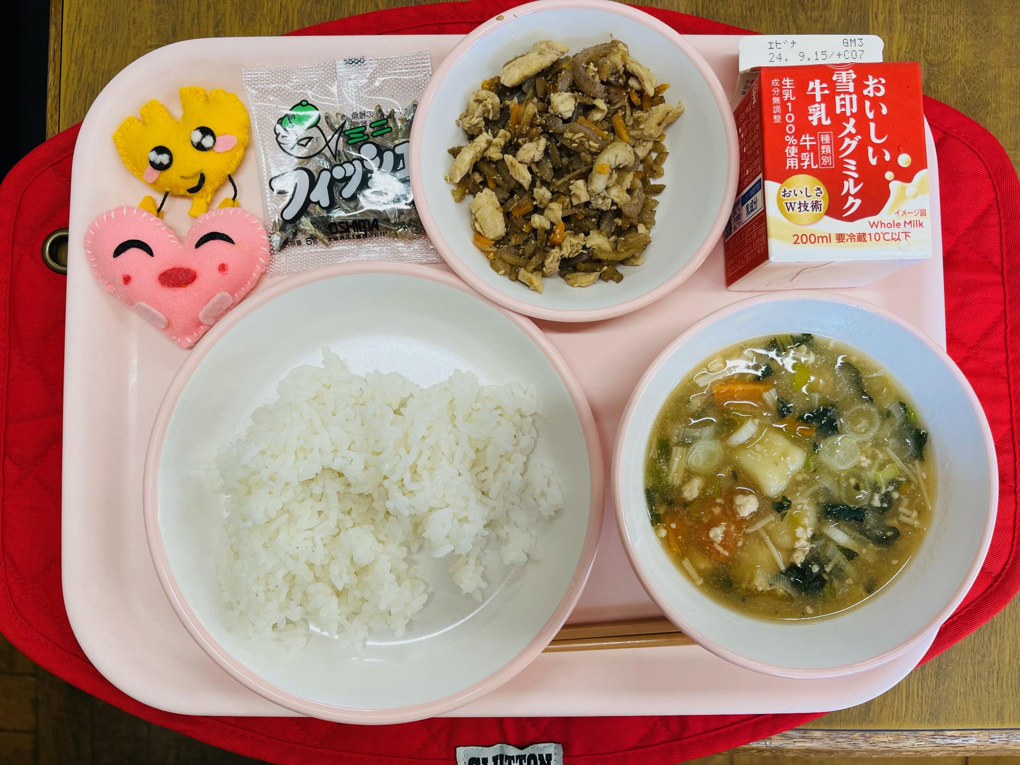 ピンクのトレーに給食が右上から反時計回りで牛乳、鶏きんぴら、味付け小魚、ハートの形をした黄色とピンク色の布製マスコット、ごはん、いももち汁と並べられている写真