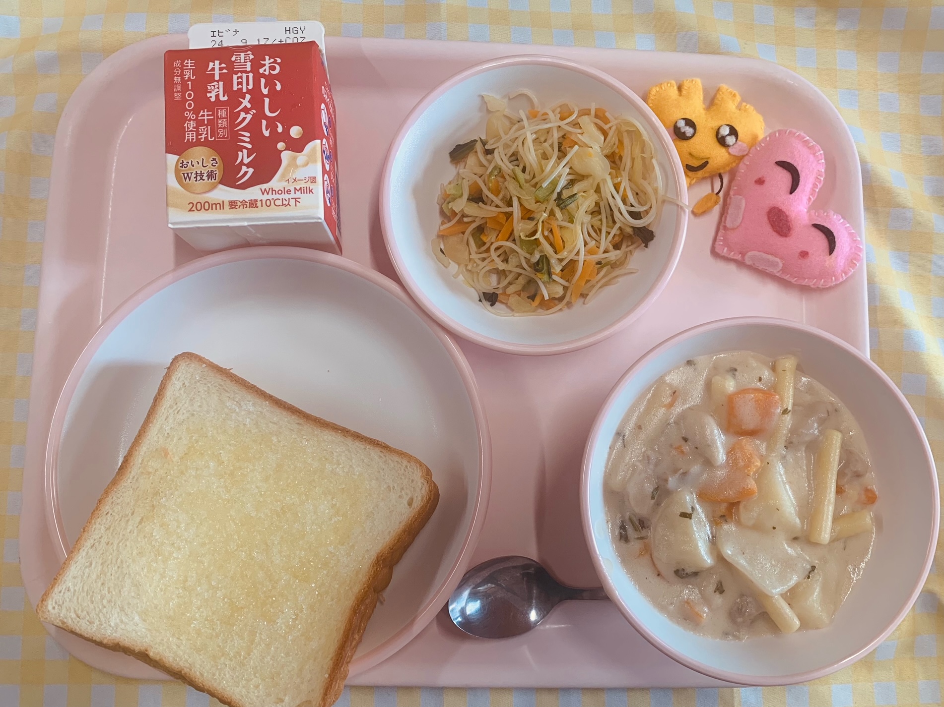 ピンクのトレーに給食が右上から反時計回りでハートの形をした黄色とピンク色の布製マスコット、ビーフンソテー、牛乳、シュガートースト、鶏肉のクリーム煮と並べられている写真