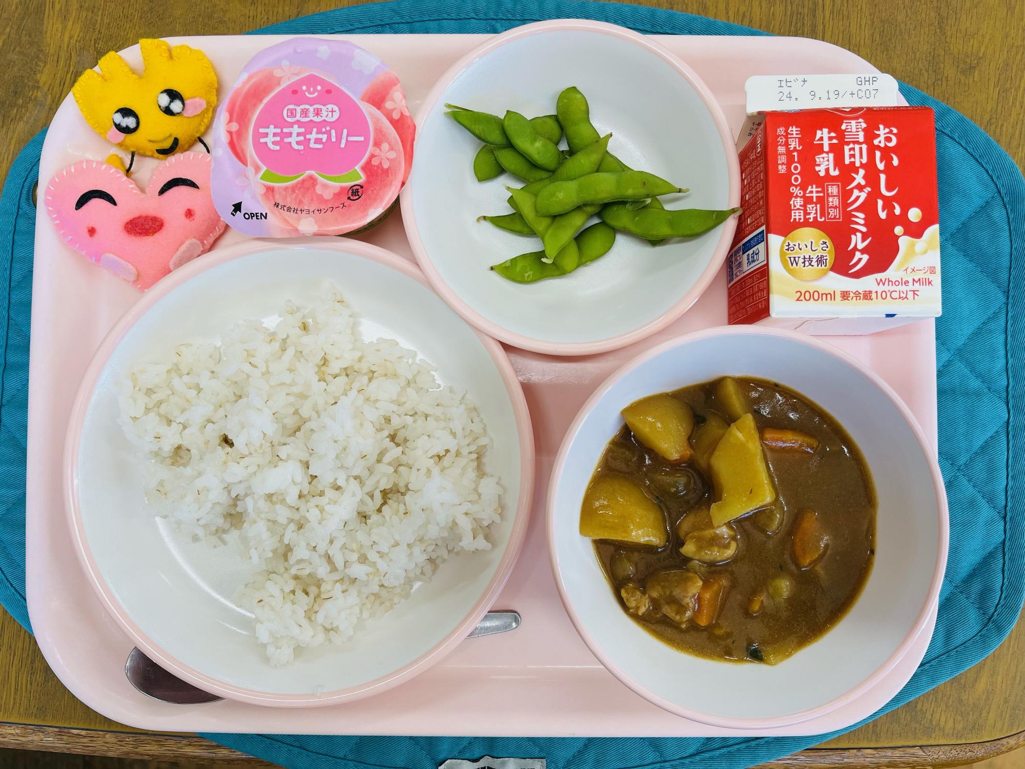 ピンクのトレーに給食が右上から反時計回りで牛乳、枝豆、桃ゼリー、ハートの形をした黄色とピンク色の布製マスコット、ごはん、チキンカレーと並べられている写真