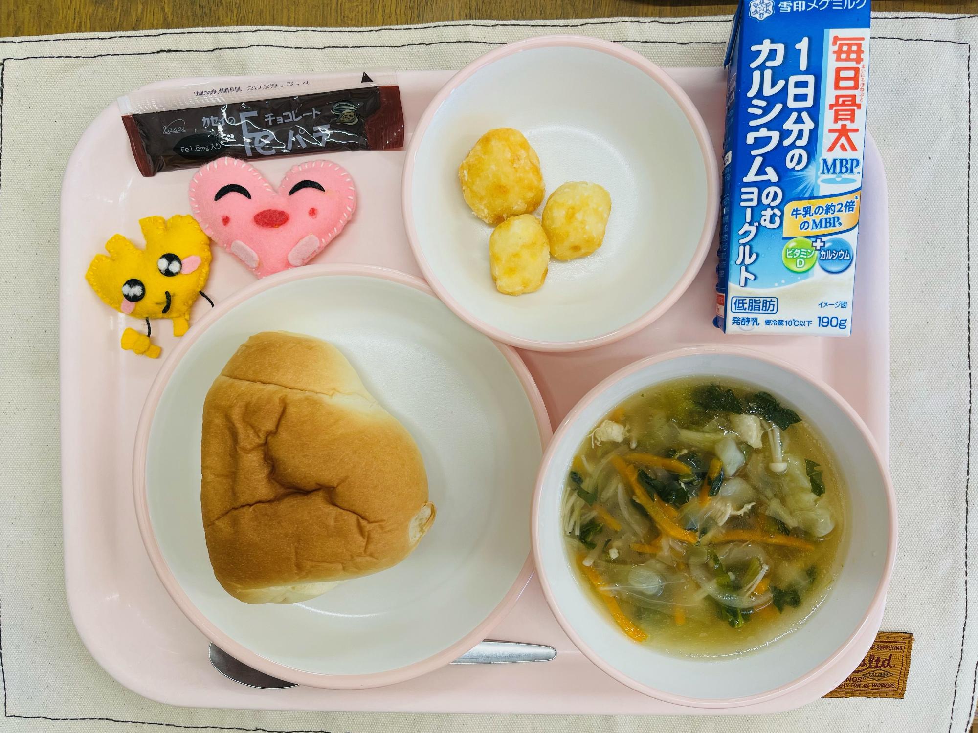 ピンクのトレーに給食が右上から反時計回りでのむヨーグルト、じゃがまる、チョコパテ、ハートの形をした黄色とピンク色の布製マスコット、ロールパン、コンソメスープと並べられている写真