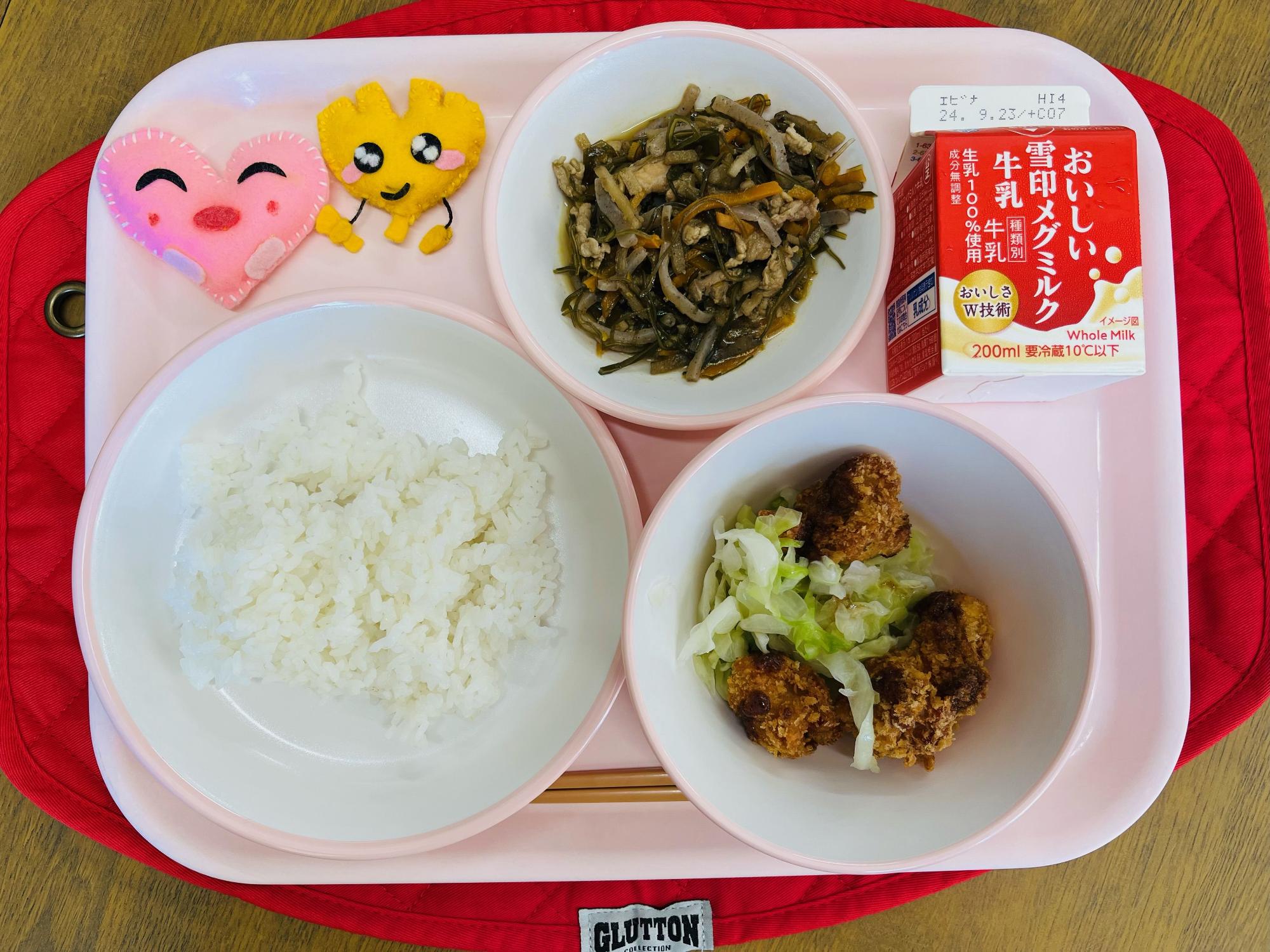 ピンクのトレーに給食が右上から反時計回りで牛乳、昆布のきんぴら、ハートの形をした黄色とピンク色の布製マスコット、ごはん、キャベツソテーとチキンカツが並べられている写真