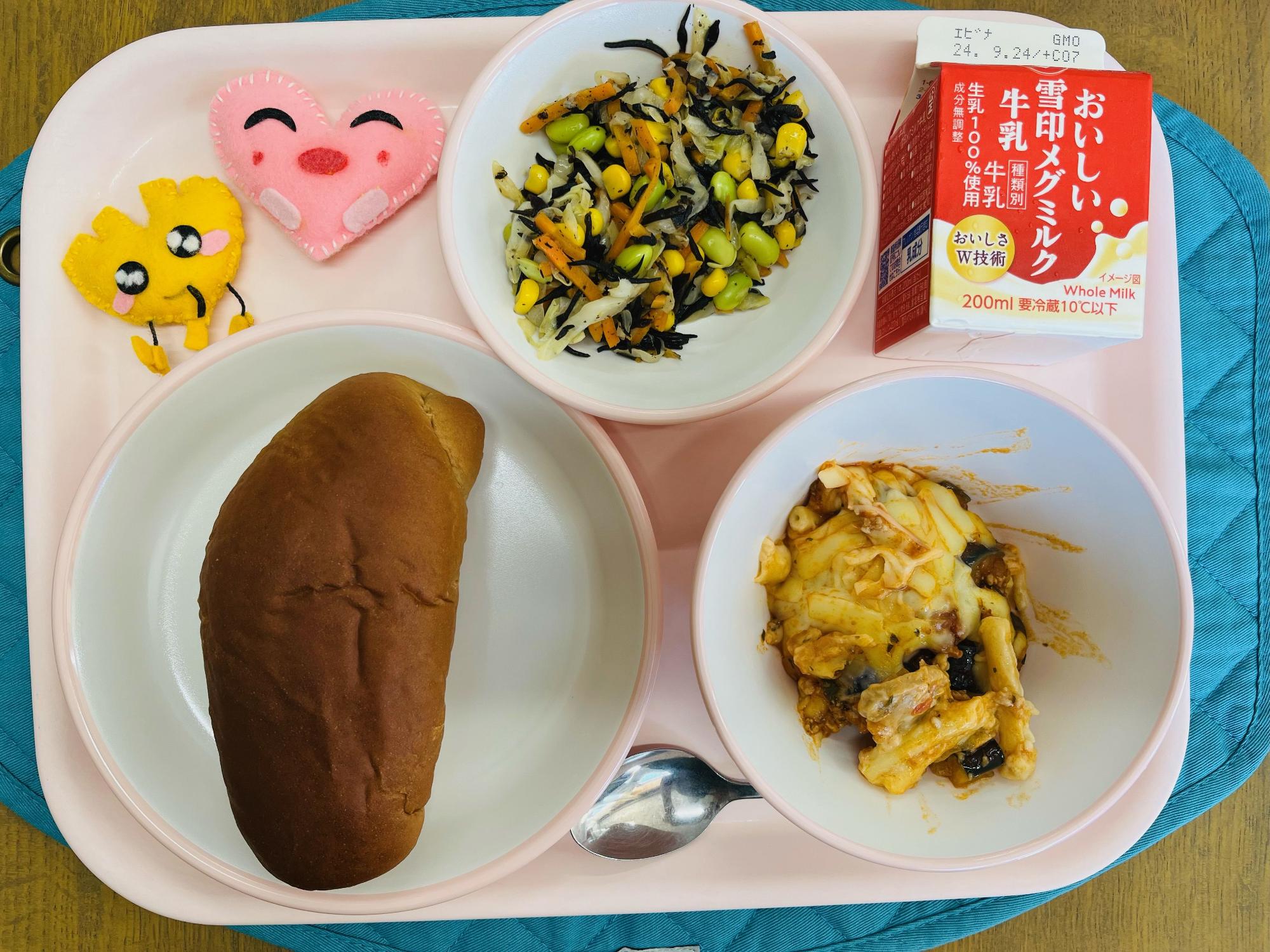 ピンクのトレーに給食が右上から反時計回りで牛乳、ひじきのサラダ、ハートの形をした黄色とピンク色の布製マスコット、くろパン、なすのミートグラタンと並べられている写真
