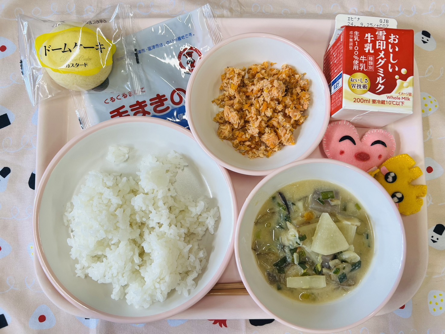 ピンクのトレーに給食が右上から反時計回りで牛乳、さけそぼろ、手まきのり、ドームケーキ、ごはん、ごじる、ハートの形をした黄色とピンク色の布製マスコットと並べられている写真