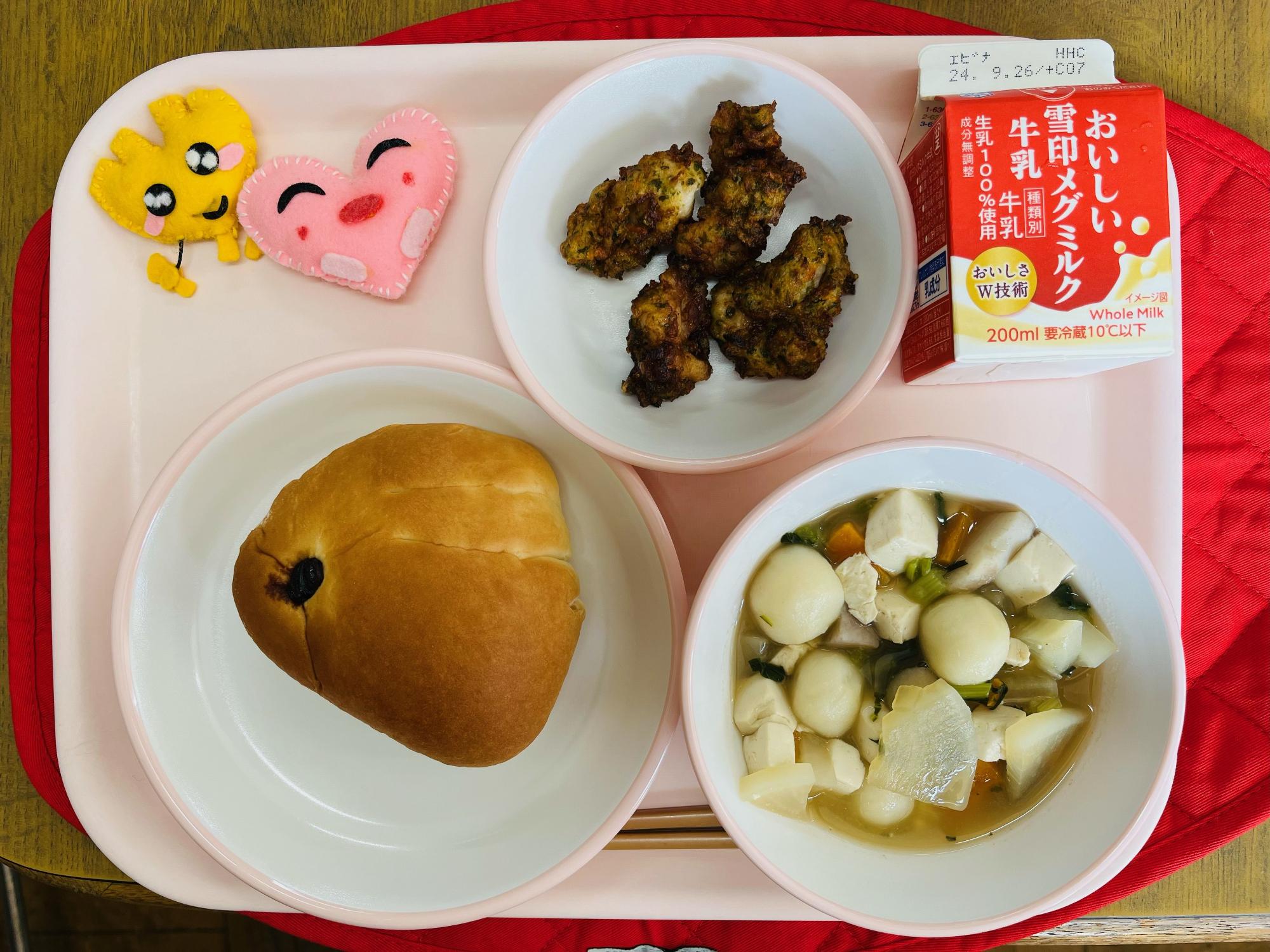 ピンクのトレーに給食が右上から反時計回りで牛乳、鶏肉のころも揚げ、ハートの形をした黄色とピンク色の布製マスコット、うさぎパン、月見汁と並べられている写真
