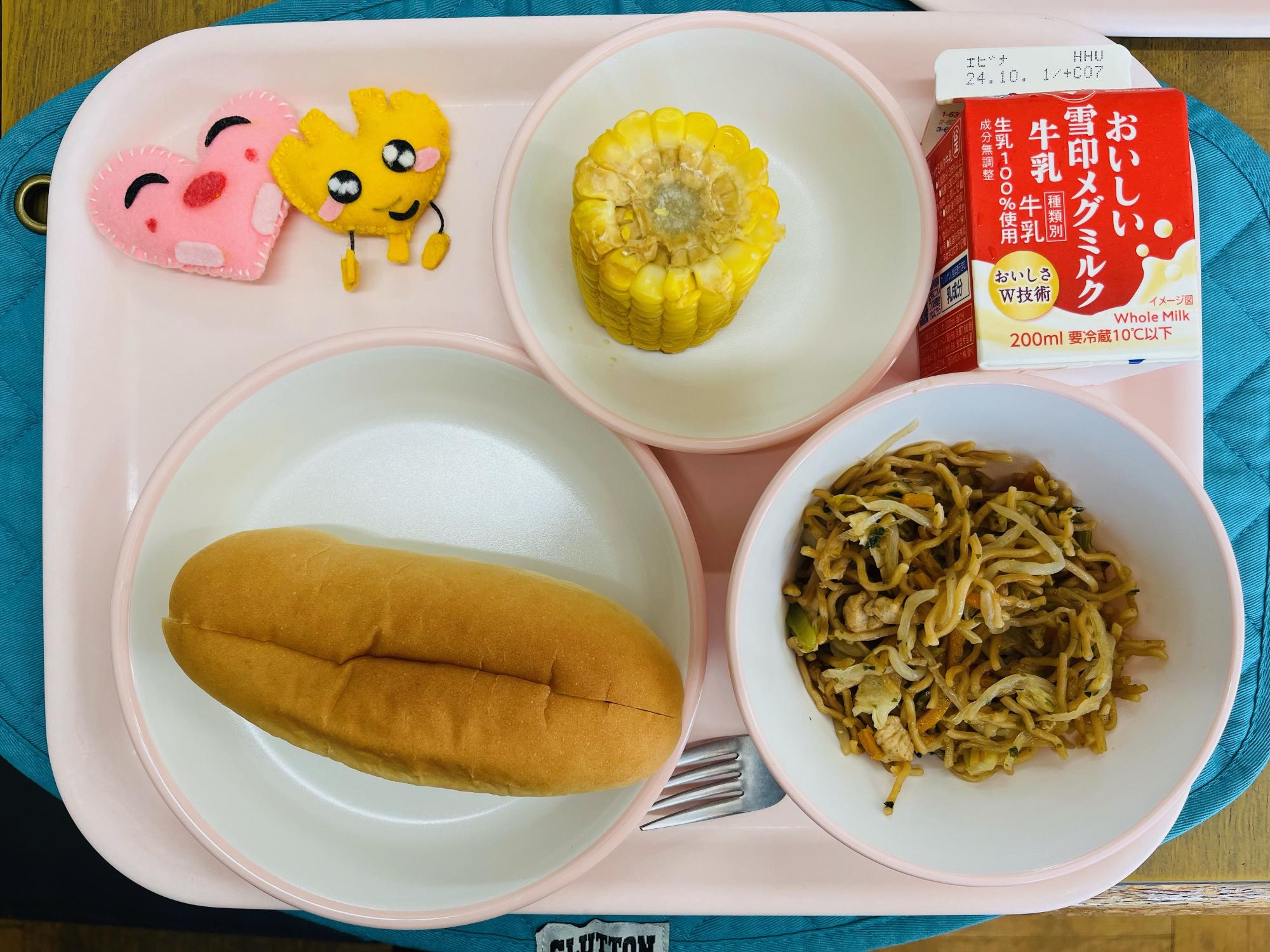 ピンクのトレーに給食が右上から反時計回りで牛乳、焼きとうもろこし、ハートの形をした黄色とピンク色の布製マスコット、サンドパン、やきそばと並べられている写真