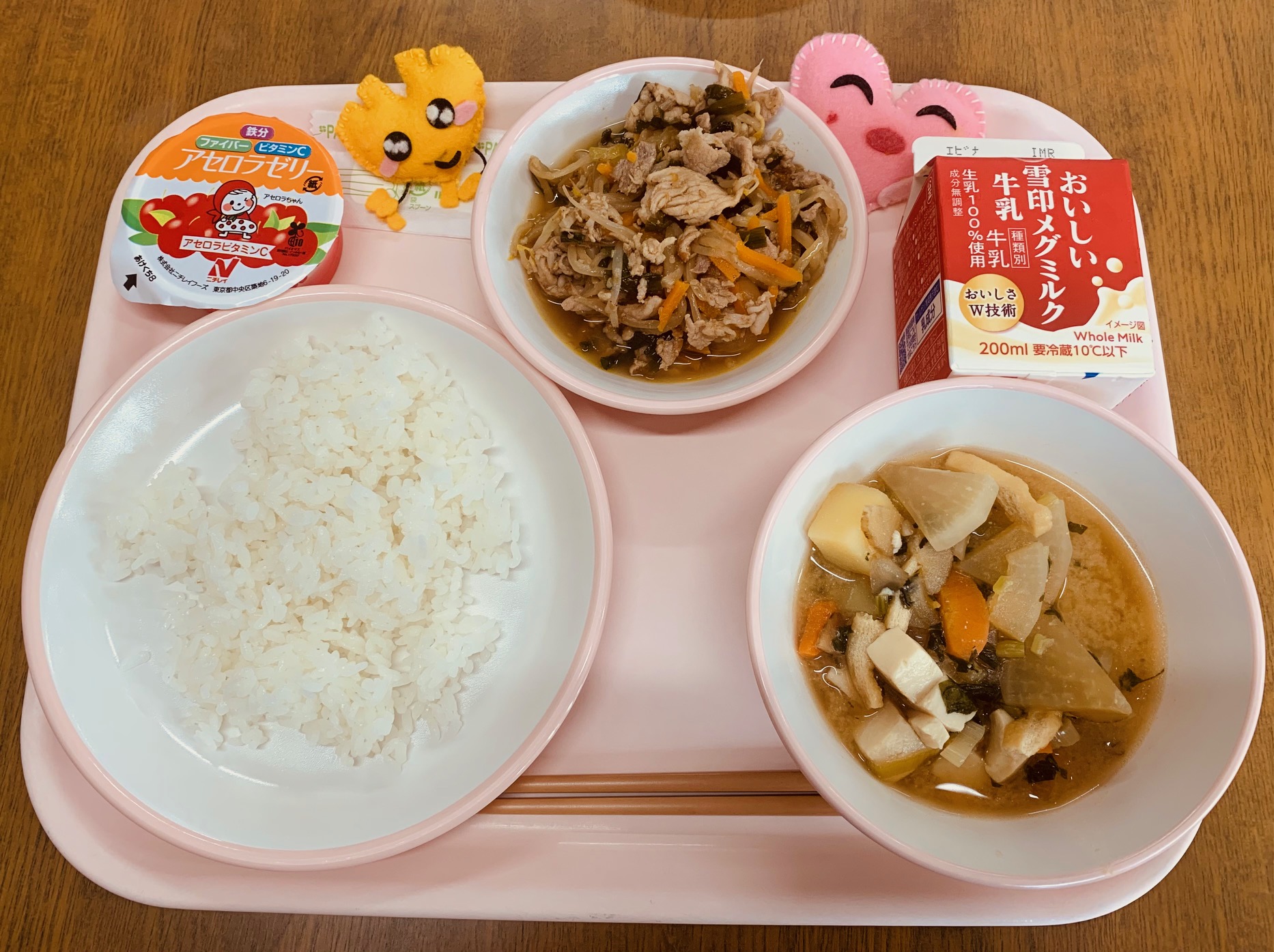 ピンクのトレーに給食が右上から反時計回りで牛乳、野菜入り焼肉、ハートの形をした黄色とピンク色の布製マスコット、アセロラゼリー、ごはん、味噌けんちん汁と並べられている写真