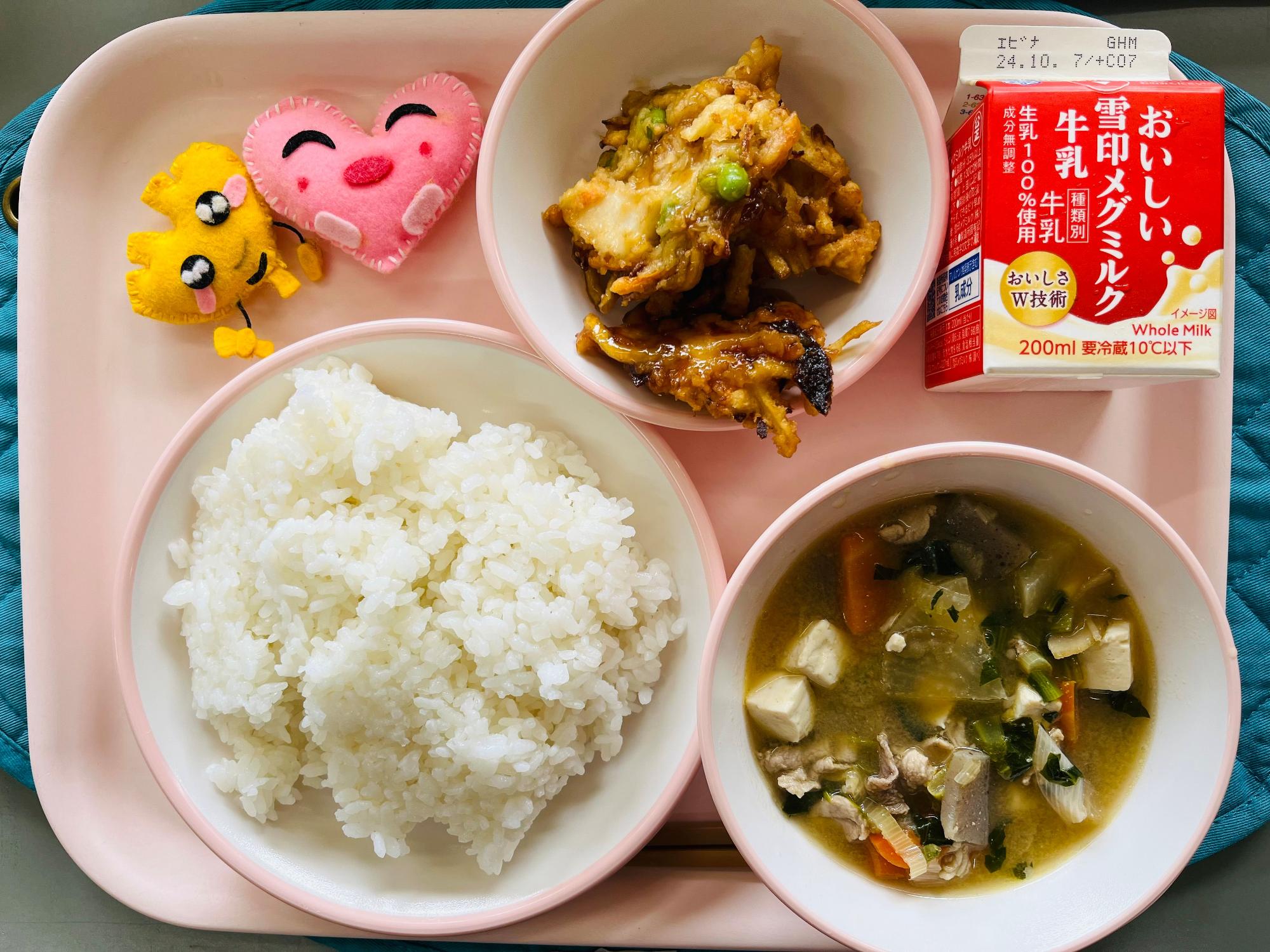 ピンクのトレーに給食が右上から反時計回りで牛乳、かきあげ、ハートの形をした黄色とピンク色の布製マスコット、ごはん、あっさり豚汁と並べられている写真