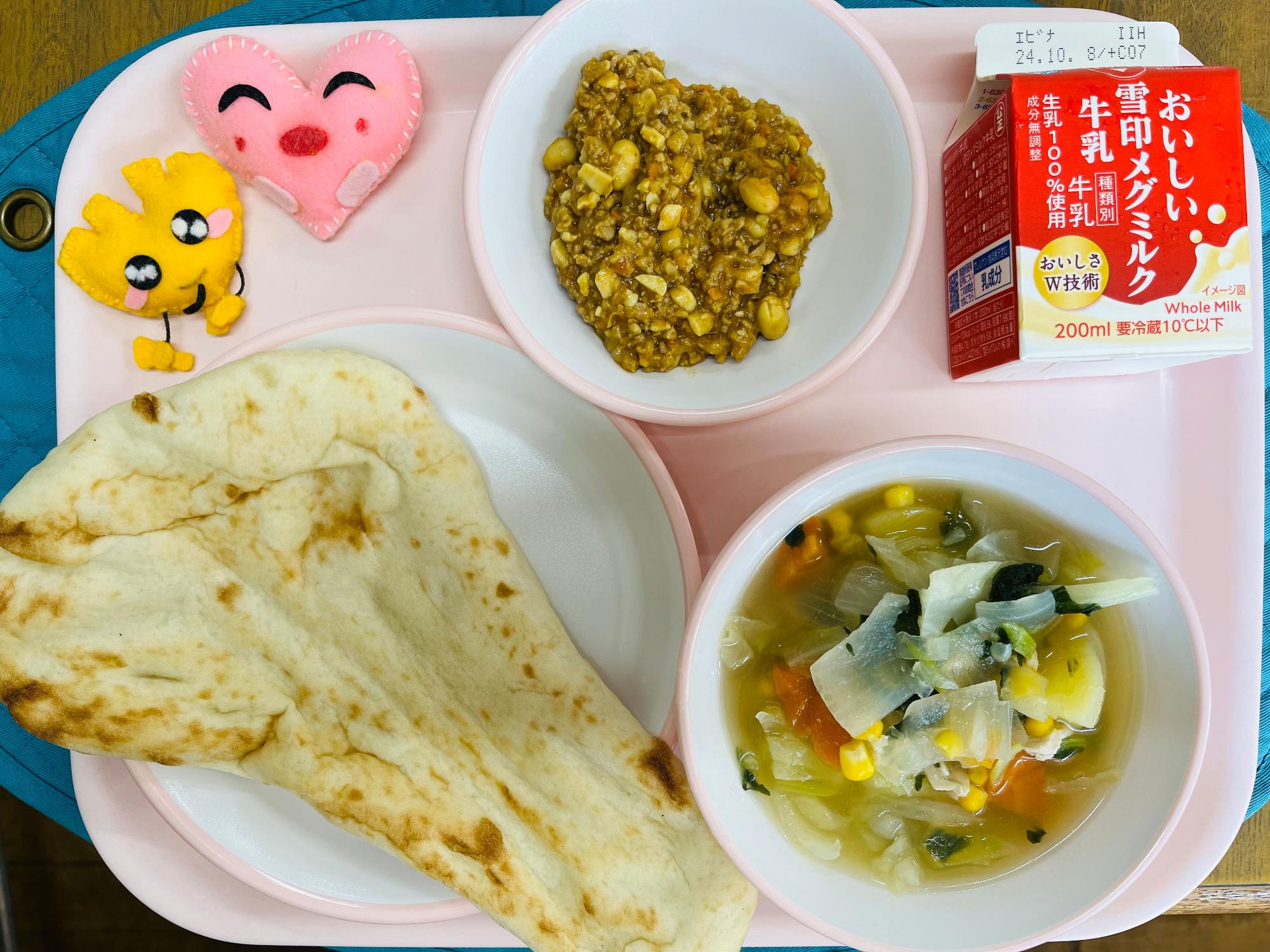 ピンクのトレーに給食が右上から反時計回りで牛乳、ドライカレー、ハートの形をした黄色とピンク色の布製マスコット、ナン、野菜のスープ煮と並べられている写真