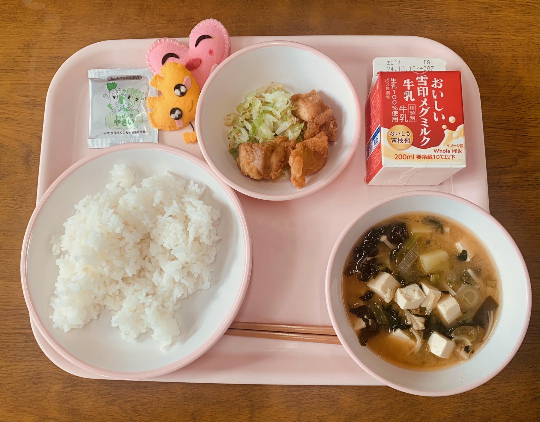 ピンクのトレーに給食が右上から反時計回りで牛乳、やみつきキャベツと鶏肉のからあげ、ハートの形をした黄色とピンク色の布製マスコット、野菜ふりかけ、ごはん、味噌汁と並べられている写真