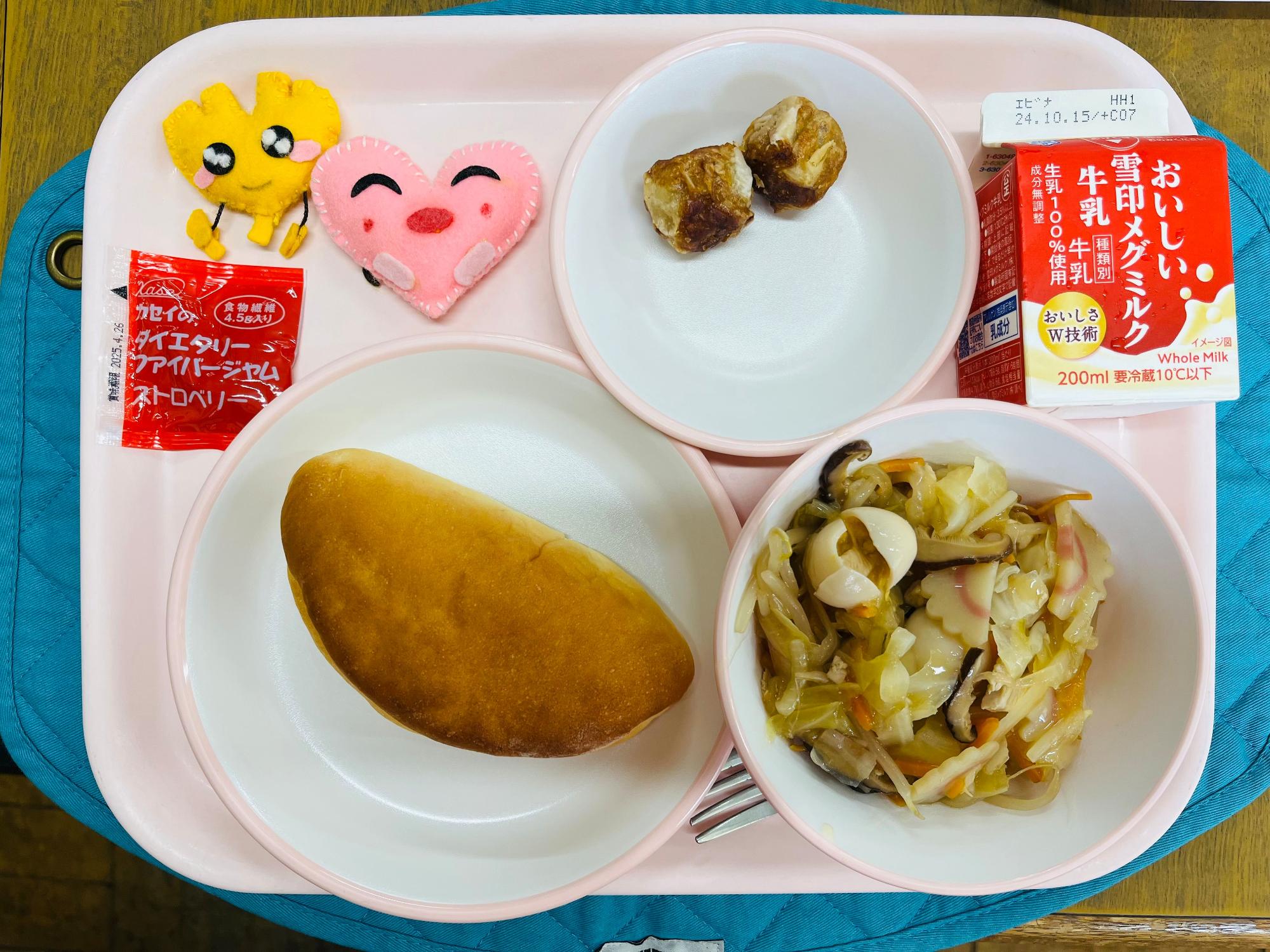ピンクのトレーに給食が右上から反時計回りで牛乳、あげシュウマイ、ハートの形をした黄色とピンク色の布製マスコット、いちごジャム、サンドパン、やきそばあんかけと並べられている写真