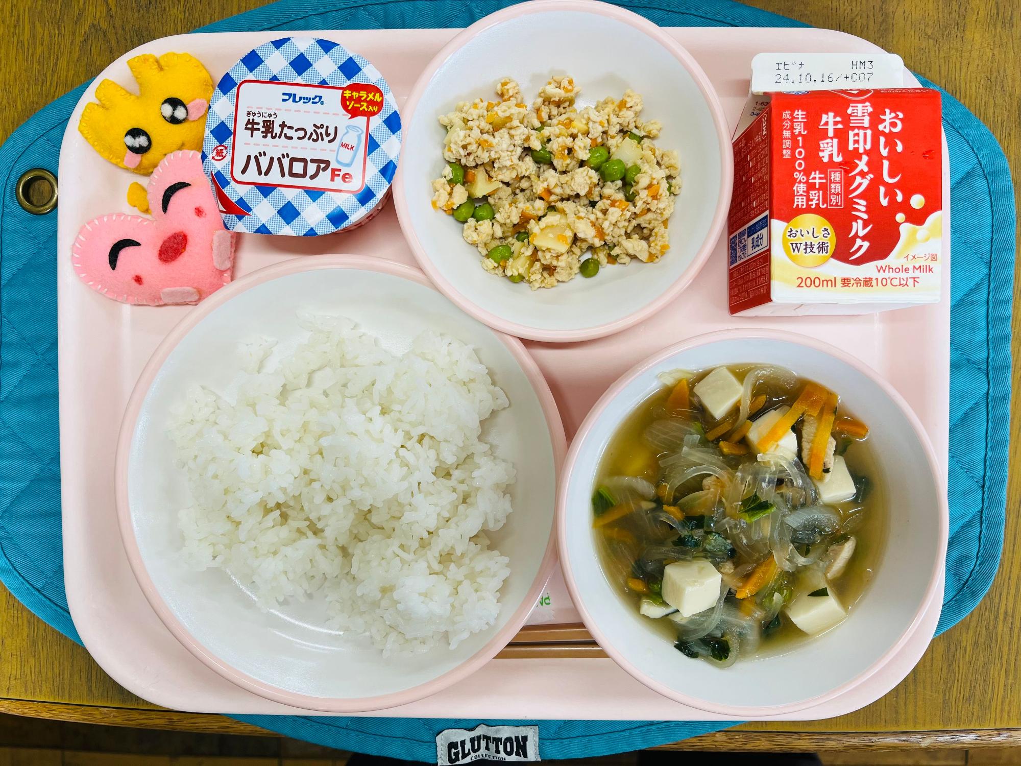 ピンクのトレーに給食が右上から反時計回りで牛乳、鶏そぼろ、ババロア、ハートの形をした黄色とピンク色の布製マスコット、ごはん、春雨スープと並べられている写真