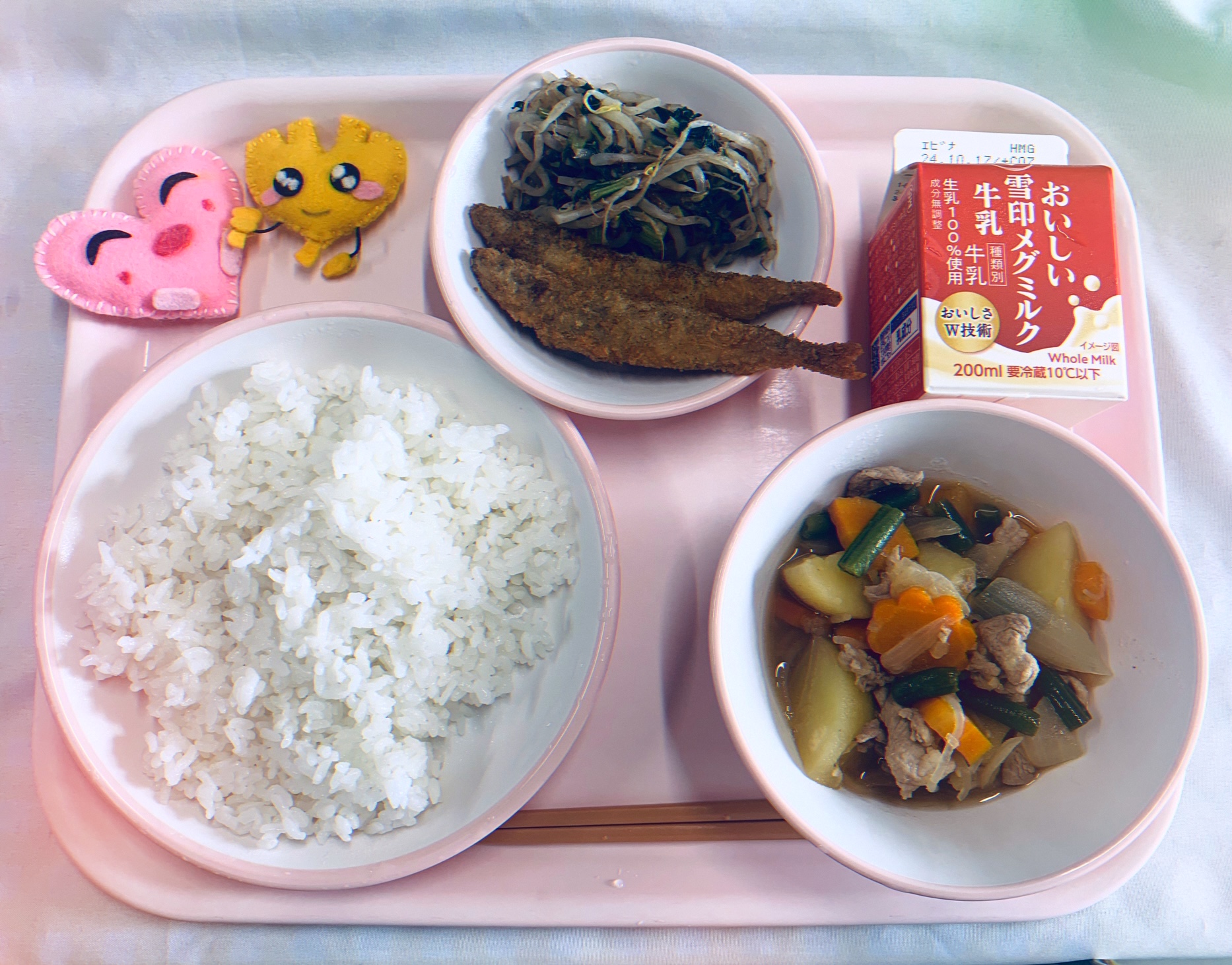 ピンクのトレーに給食が右上から反時計回りで牛乳、ししゃもフライともやしのおかかあえ、ハートの形をした黄色とピンク色の布製マスコット、ごはん、肉じゃがと並べられている写真