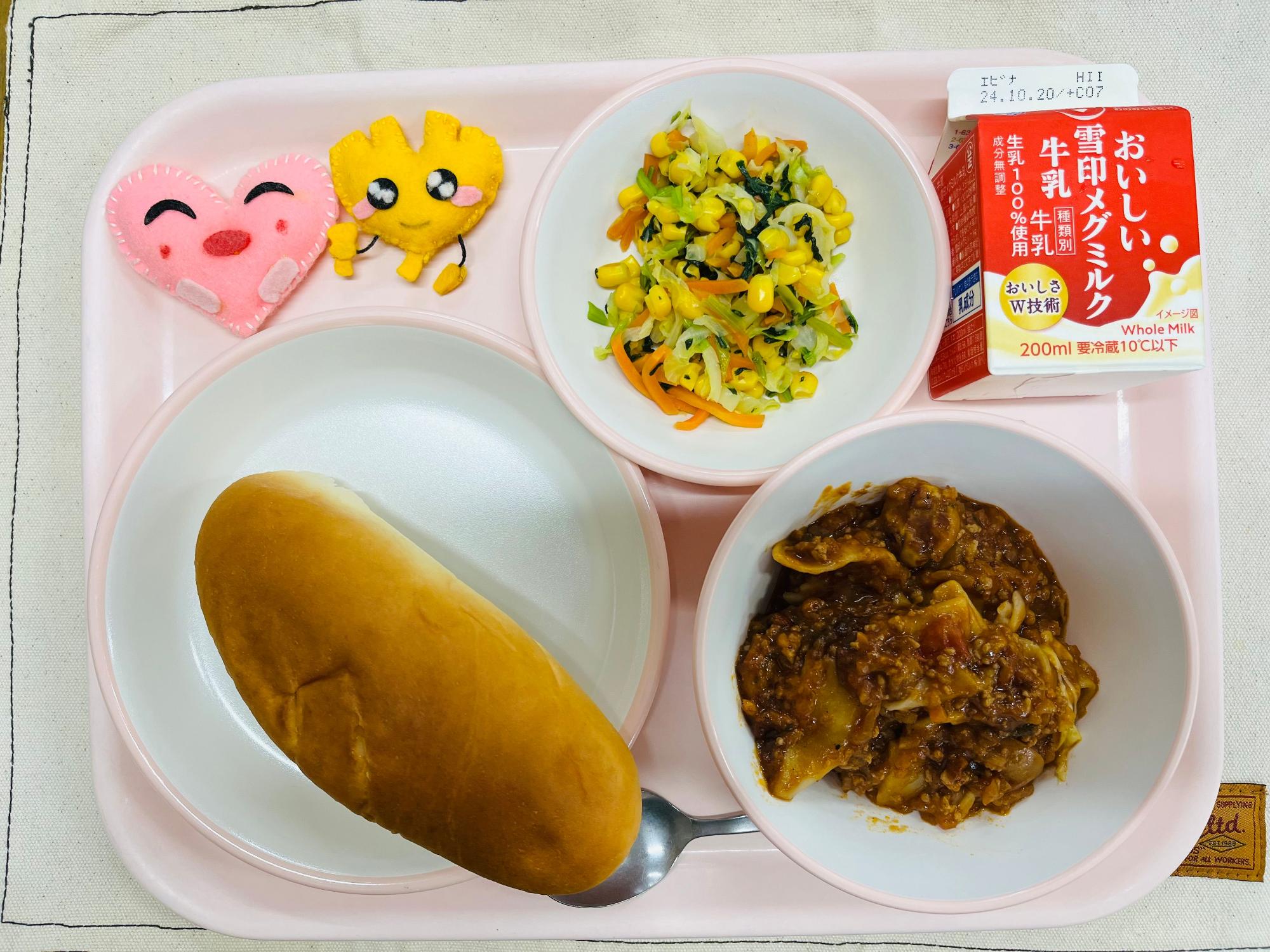 ピンクのトレーに給食が右上から反時計回りで牛乳、コーンソテー、ハートの形をした黄色とピンク色の布製マスコット、ミルクパン、ラザニアと並べられている写真