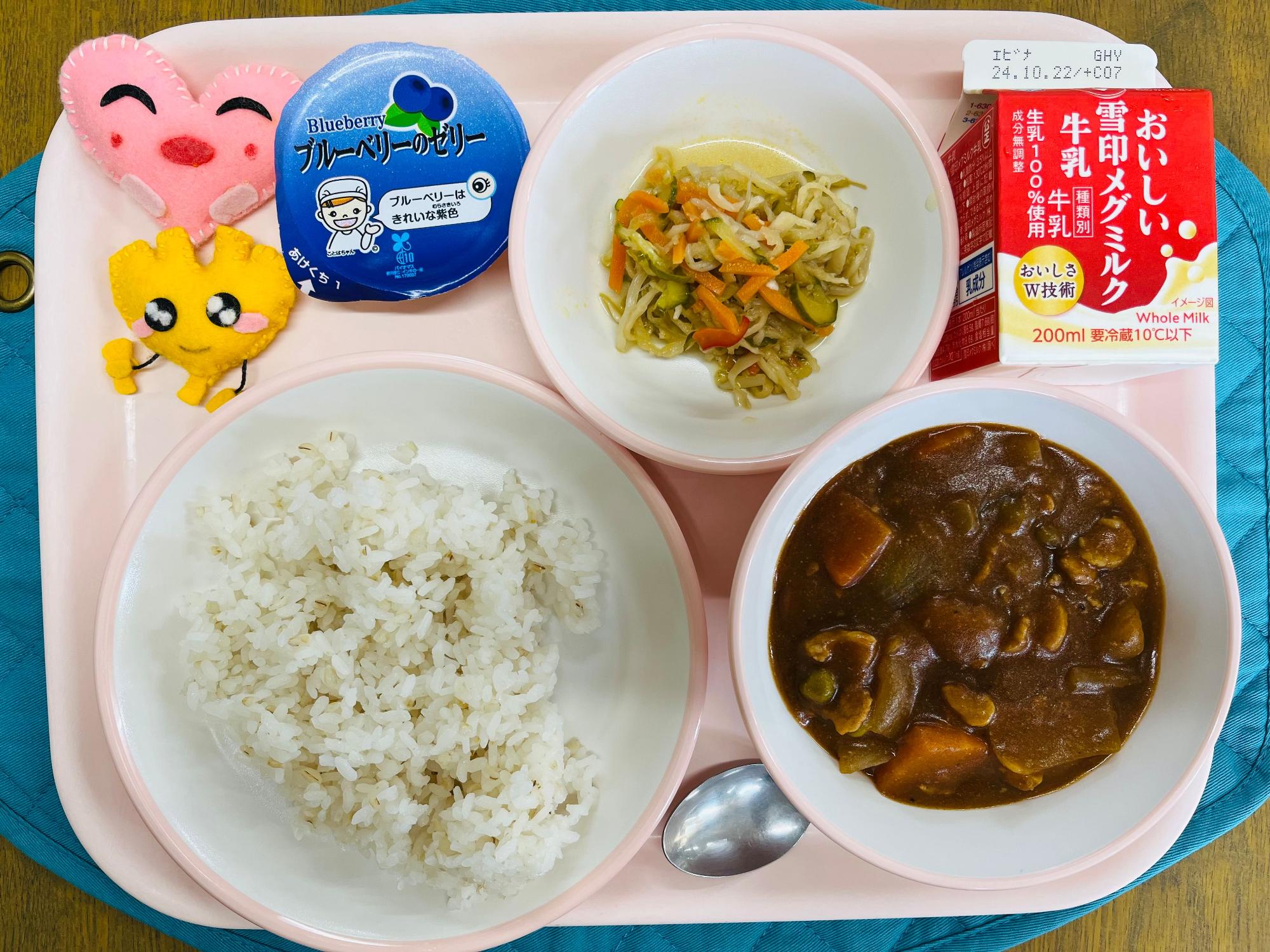 ピンクのトレーに給食が右上から反時計回りで牛乳、切り干し大根のサラダ、ブルーベリーゼリー、ハートの形をした黄色とピンク色の布製マスコット、ごはん、ハヤシライスと並べられている写真