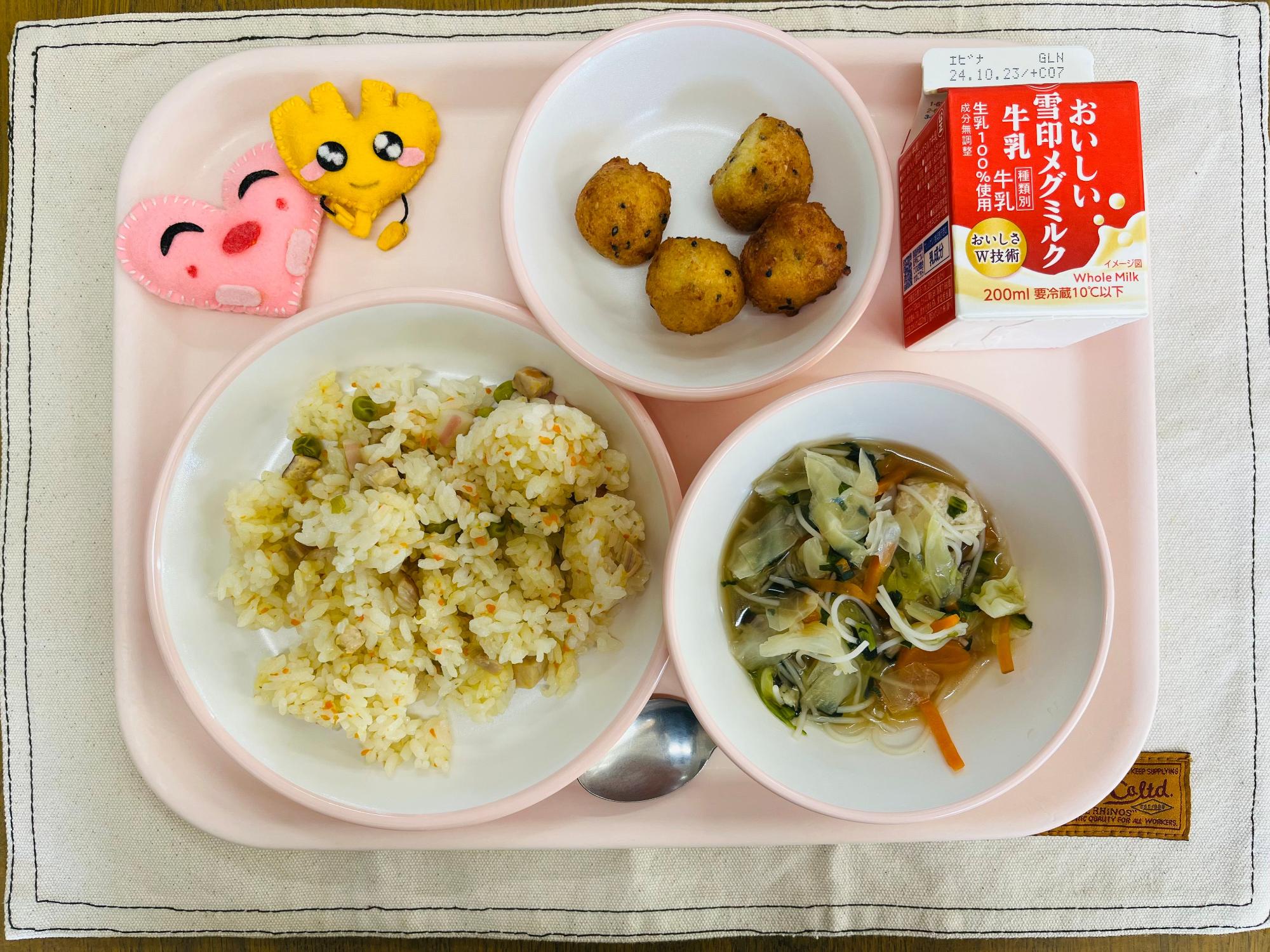 ピンクのトレーに給食が右上から反時計回りで牛乳、豆腐ドーナツ、ハートの形をした黄色とピンク色の布製マスコット、炊き込みチャーハン、鶏団子スープと並べられている写真