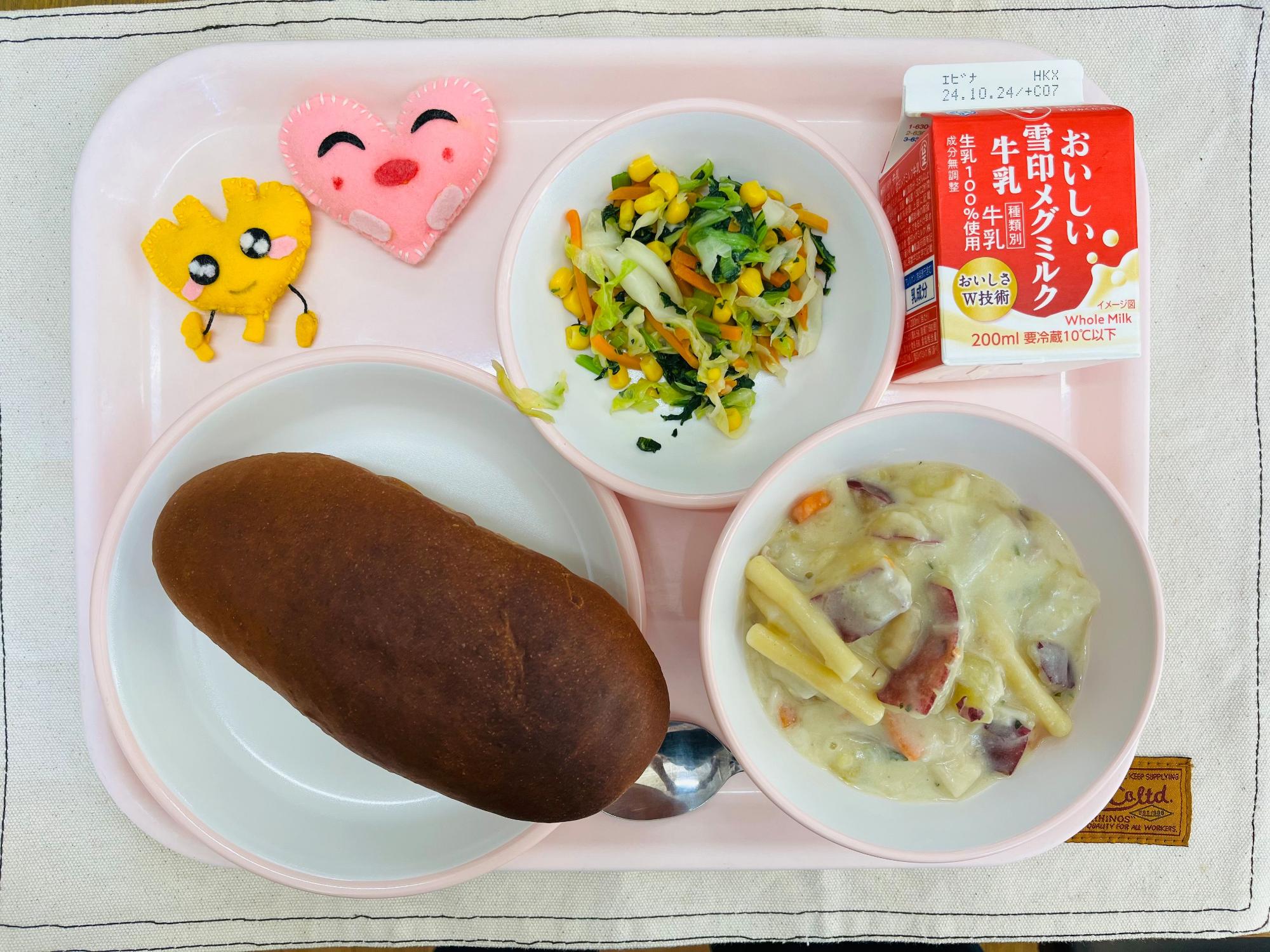 ピンクのトレーに給食が右上から反時計回りで牛乳、野菜ソテー、ハートの形をした黄色とピンク色の布製マスコット、くろパン、サツマイモのクリーム煮と並べられている写真