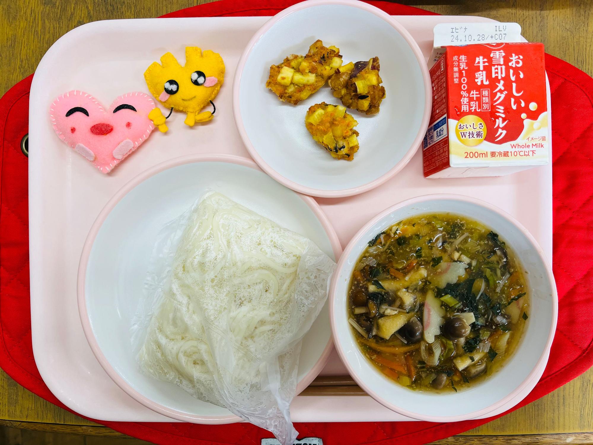 ピンクのトレーに給食が右上から反時計回りで牛乳、がんせきあげ、ハートの形をした黄色とピンク色の布製マスコット、麺、きのこうどんのスープと並べられている写真