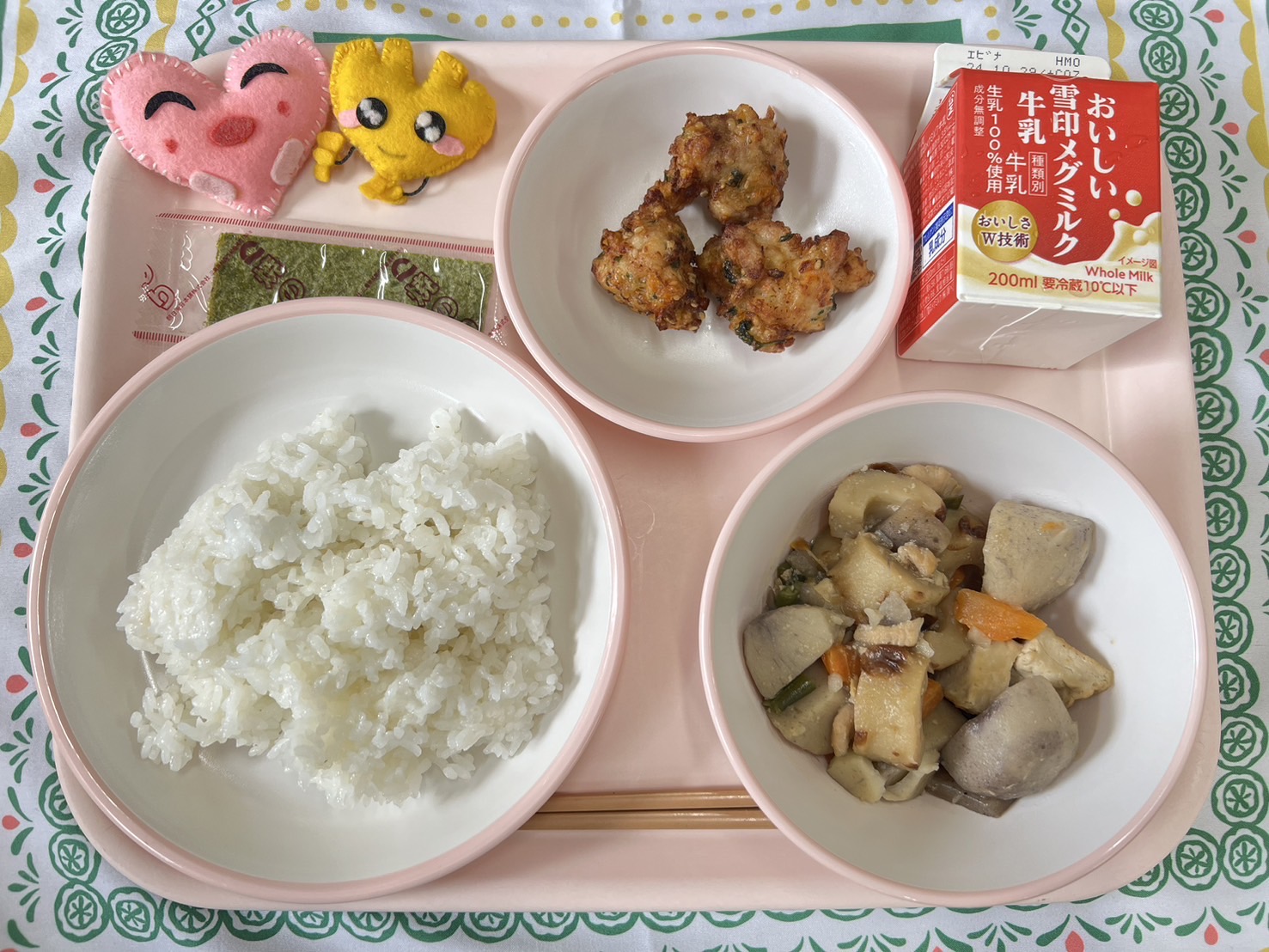 ピンクのトレーに給食が右上から反時計回りで牛乳、鶏肉の香り揚げ、ハートの形をした黄色とピンク色の布製マスコット、やきのり、ごはん、里芋と厚揚げの煮物と並べられている写真