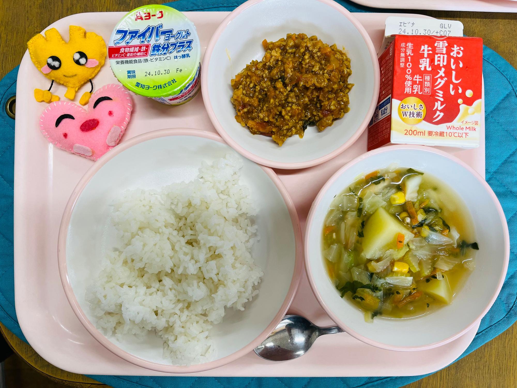 ピンクのトレーに給食が右上から反時計回りで牛乳、キーマカレー、ヨーグルト、ハートの形をした黄色とピンク色の布製マスコット、ごはん、野菜スープと並べられている写真
