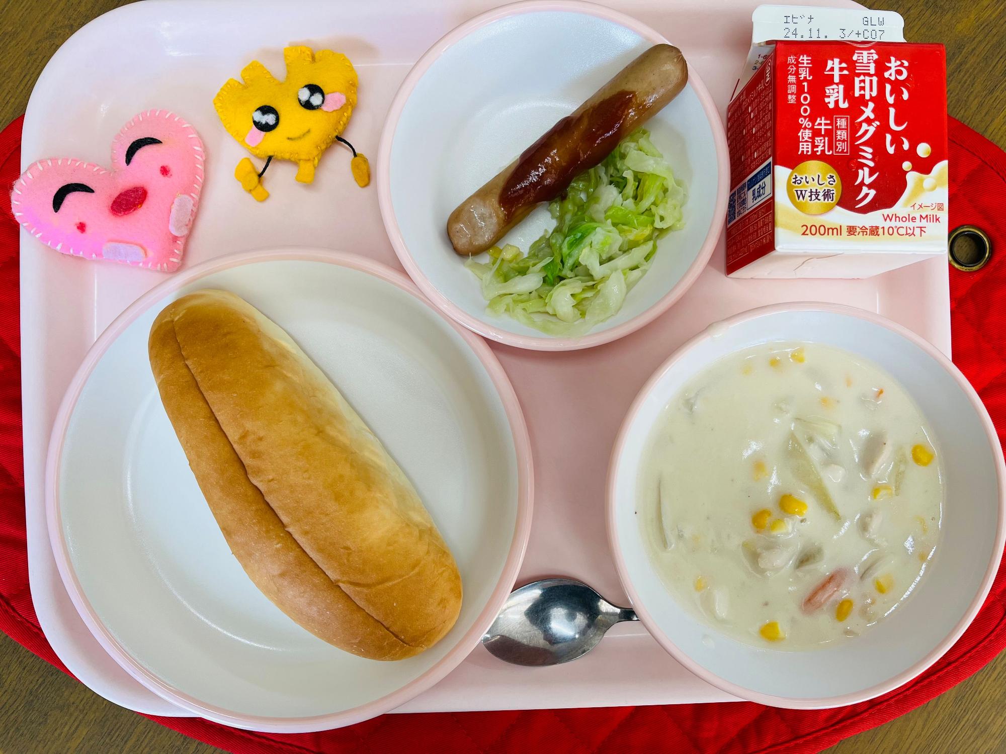 ンクのトレーに給食が右上から反時計回りで牛乳、ウインナーサンドの具、ハートの形をした黄色とピンク色の布製マスコット、サンドパン、コーンのミルクスープと並べられている写真
