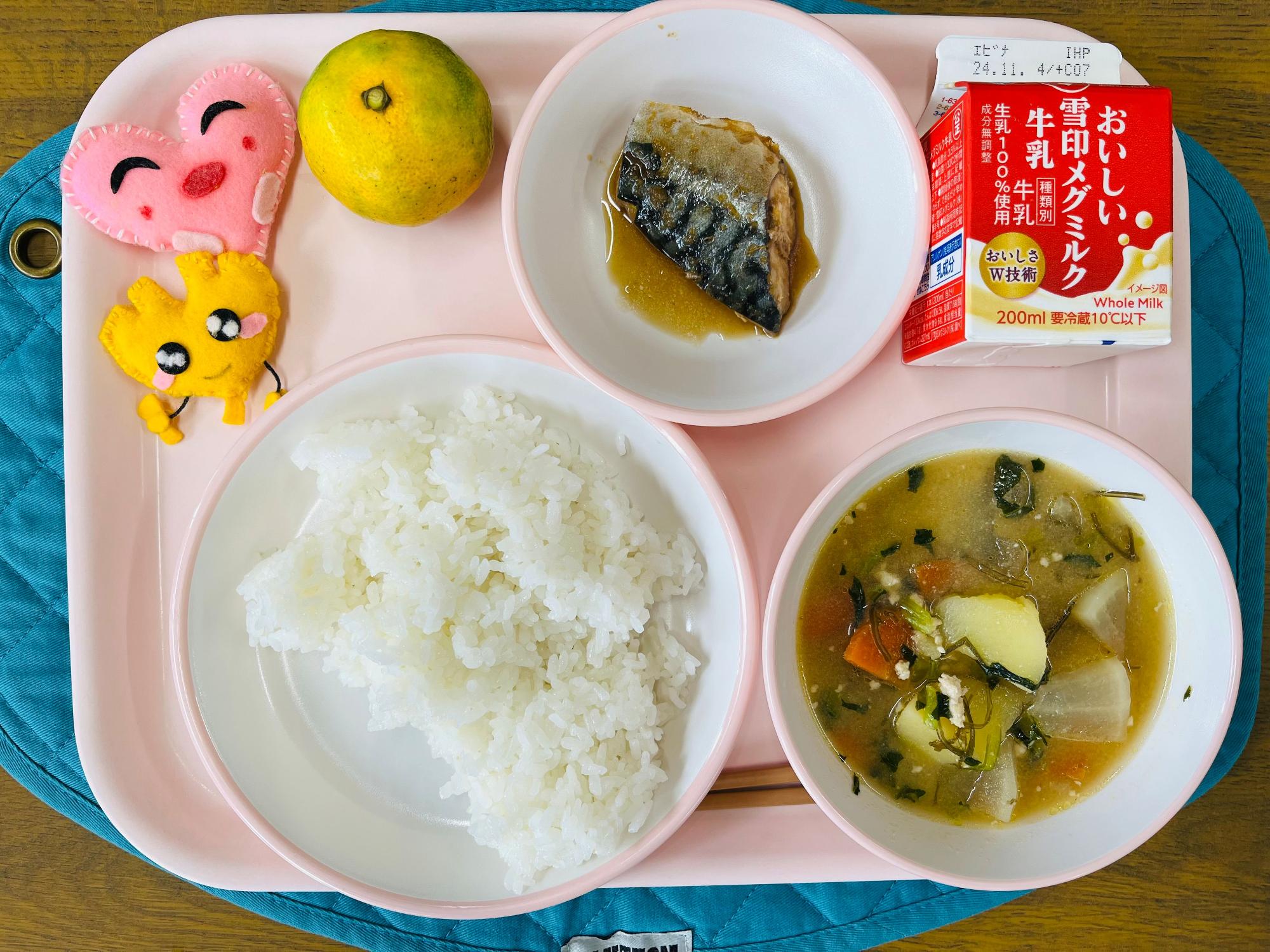 ピンクのトレーに給食が右上から反時計回りで牛乳、サバの生姜煮、みかん、ハートの形をした黄色とピンク色の布製マスコット、ごはん、じゃがこん汁と並べられている写真