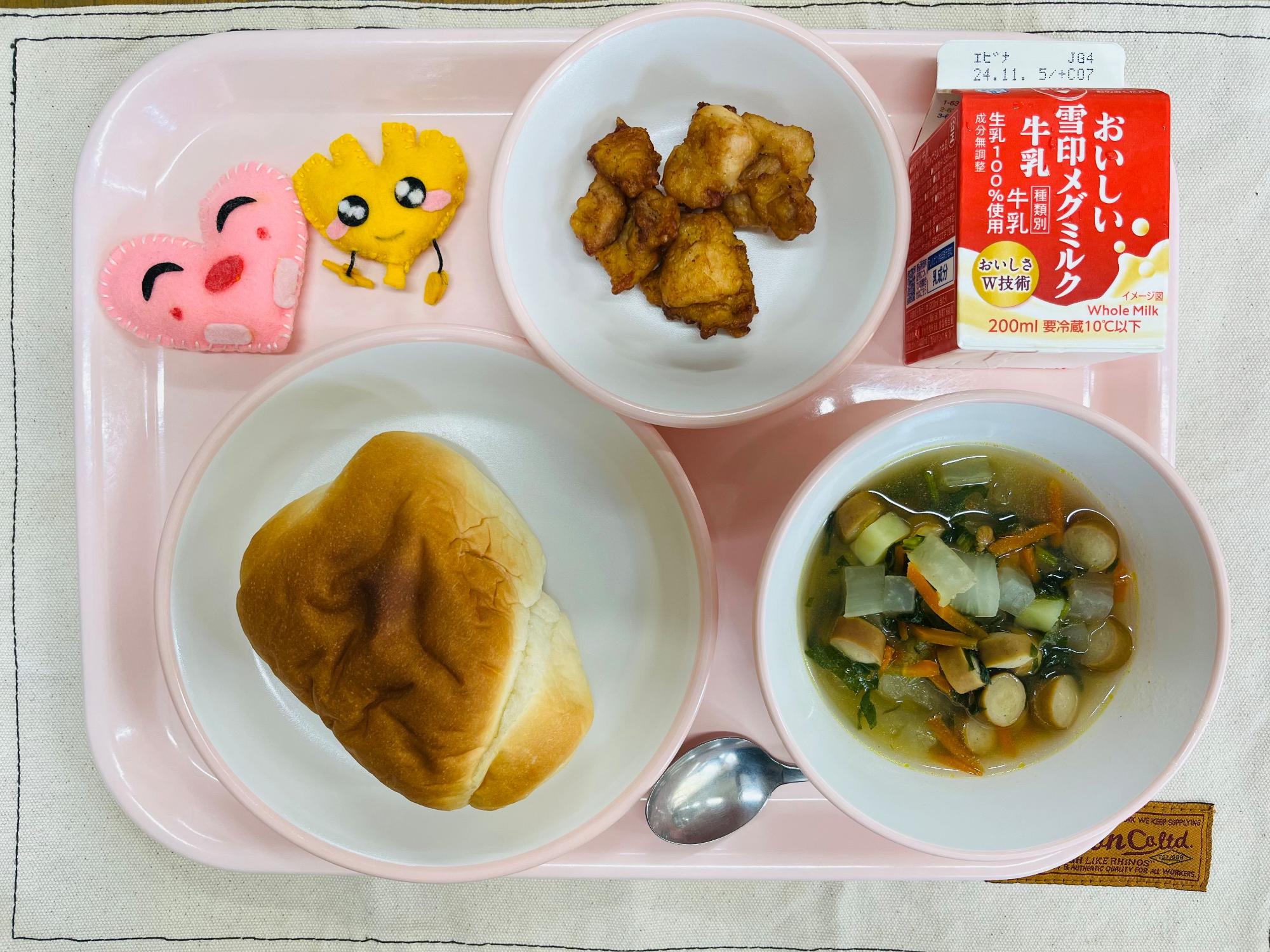ピンクのトレーに給食が右上から反時計回りで牛乳、鶏肉のフリッター、ハートの形をした黄色とピンク色の布製マスコット、ロールパン、大根のカレースープと並べられている写真