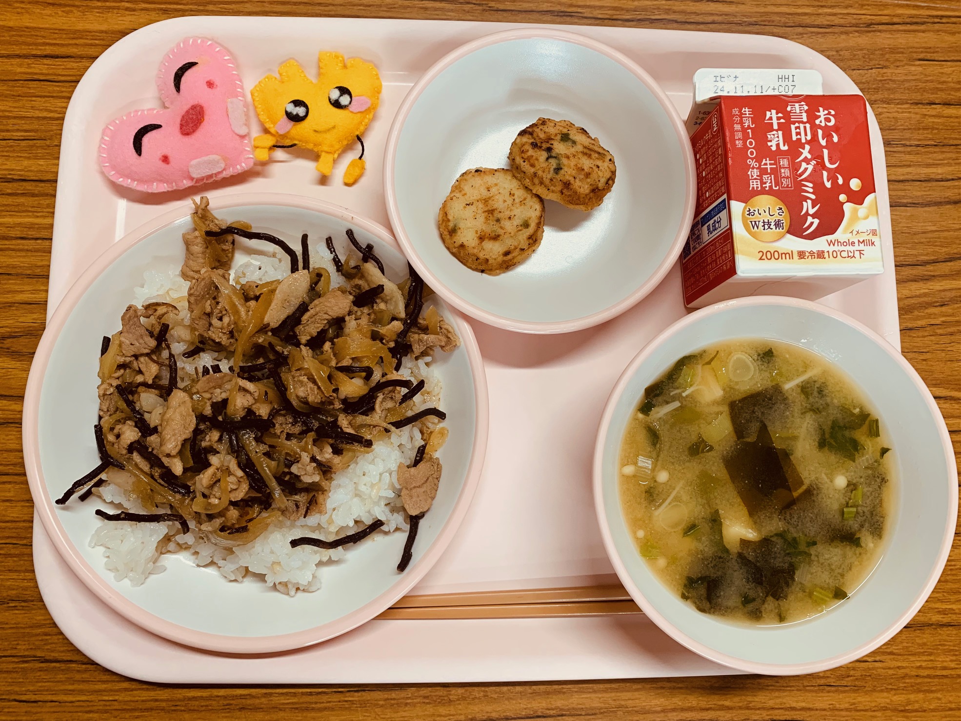 ピンクのトレーに給食が右上から反時計回りで牛乳、鶏肉と野菜のフリット、ハートの形をした黄色とピンク色の布製マスコット、豚丼、味噌汁と並べられている写真