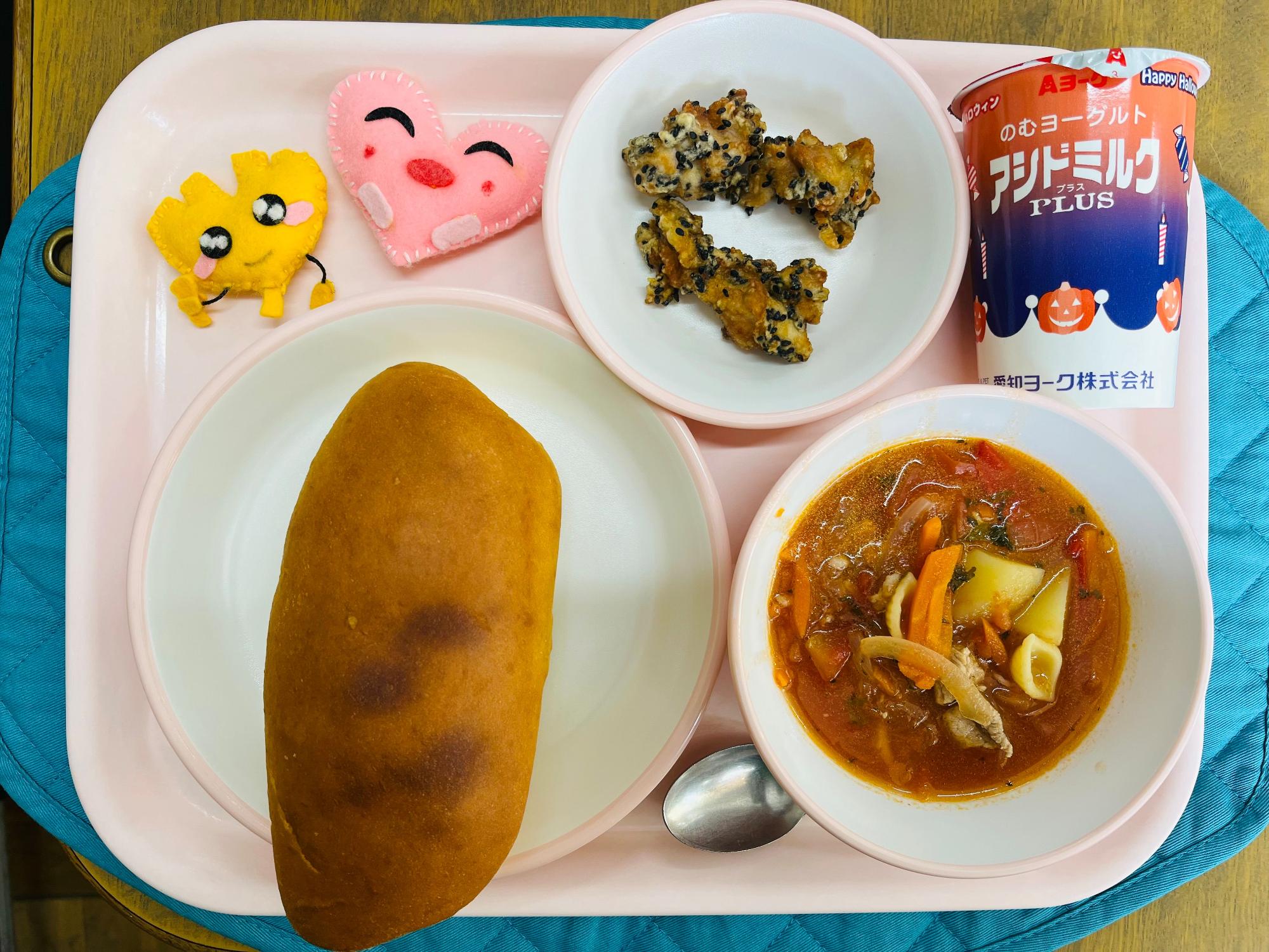 ピンクのトレーに給食が右上から反時計回りで発酵乳、黒ごまあまだれスパイシーチキン、ハートの形をした黄色とピンク色の布製マスコット、パンプキンパン、ミネストローネと並べられている写真