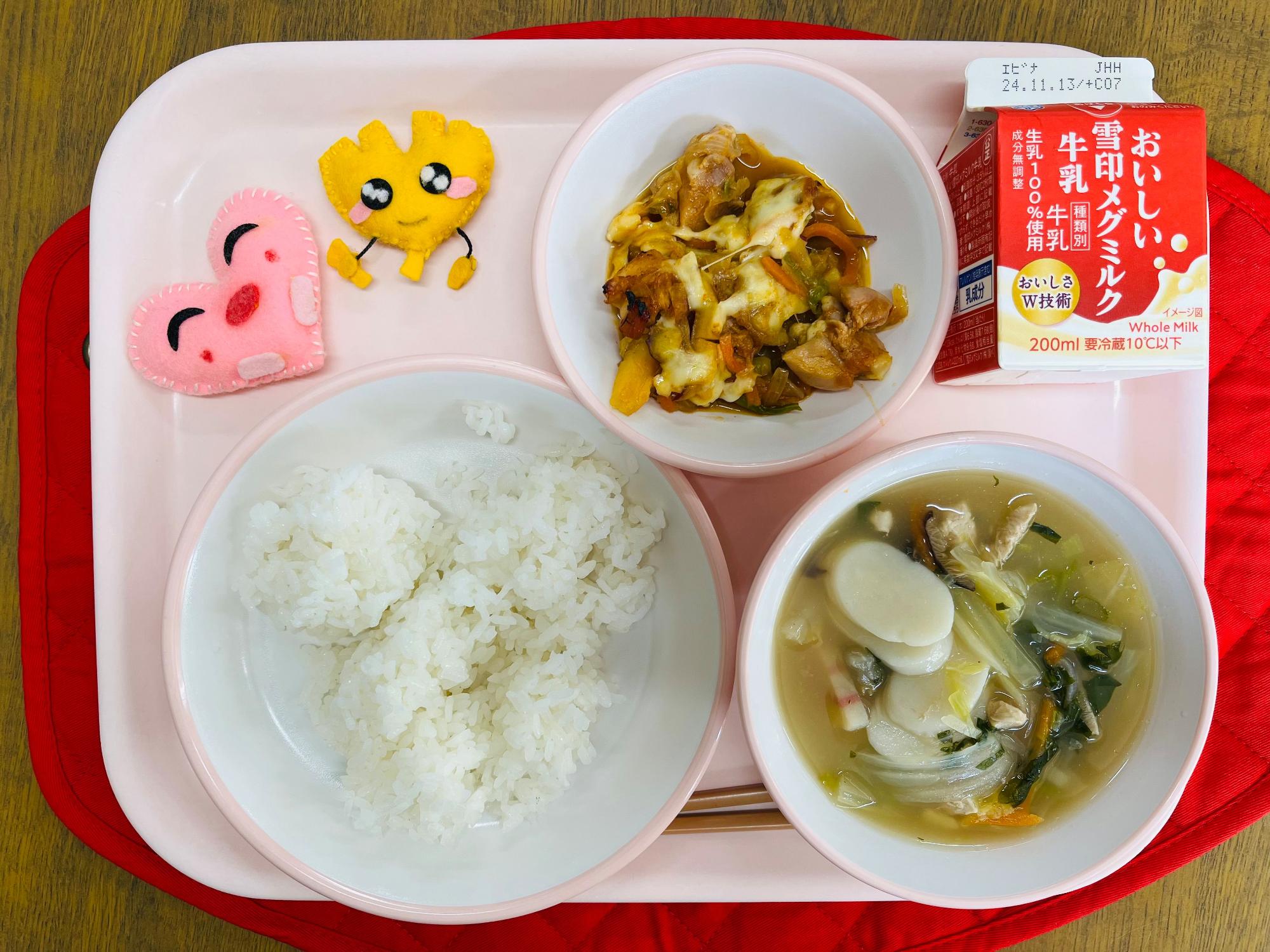 ピンクのトレーに給食が右上から反時計回りで牛乳、チーズタッカルビ、ハートの形をした黄色とピンク色の布製マスコット、ごはん、トックスープと並べられている写真