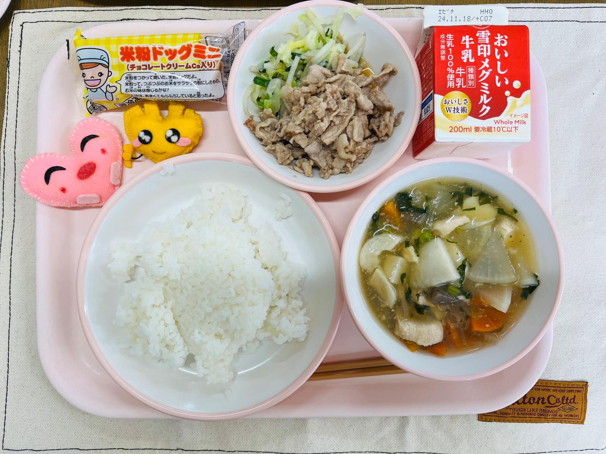 ピンクのトレーに給食が右上から反時計回りで牛乳、野菜炒めと豚肉の生姜焼き、米粉ドック、ハートの形をした黄色とピンク色の布製マスコット、ごはん、のっぺい汁と並べられている写真