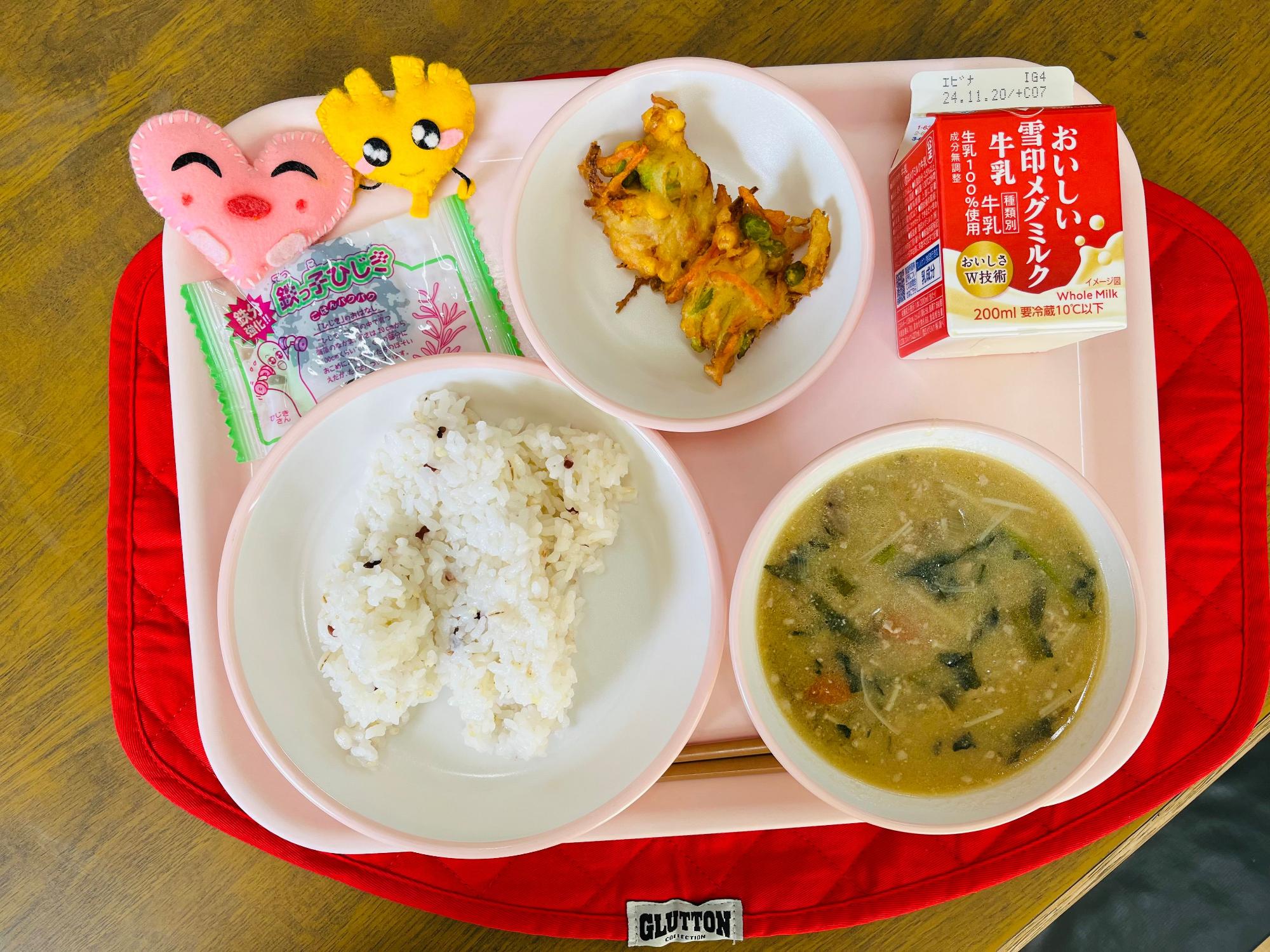 ピンクのトレーに給食が右上から反時計回りで牛乳、かみかみかきあげ、ハートの形をした黄色とピンク色の布製マスコット、佃煮、十穀米入りごはん、いももち汁と並べられている写真