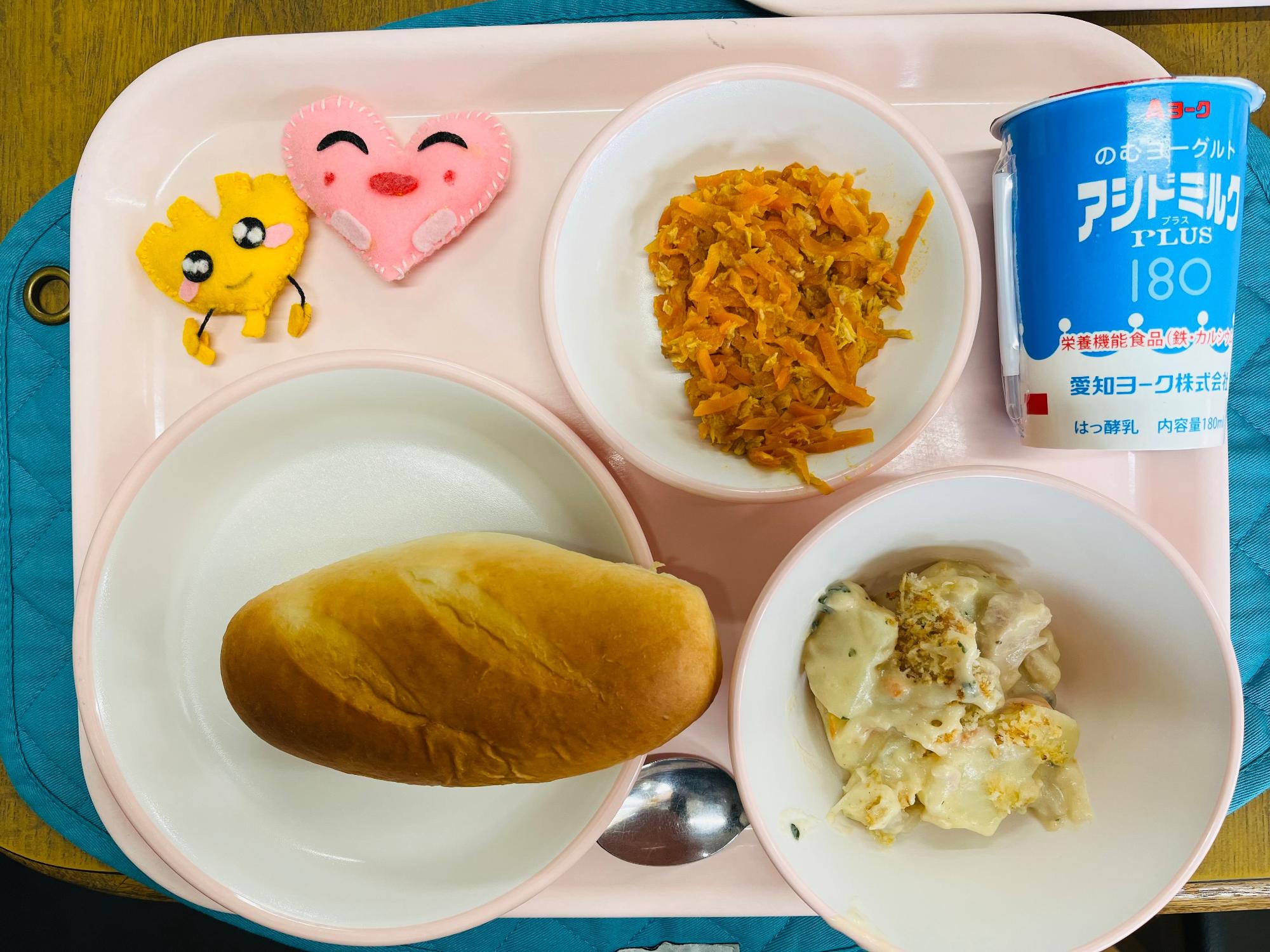 ピンクのトレーに給食が右上から反時計回りで発酵乳、にんじんシリシリ、ハートの形をした黄色とピンク色の布製マスコット、ミルクパン、秋の味覚グラタンと並べられている写真