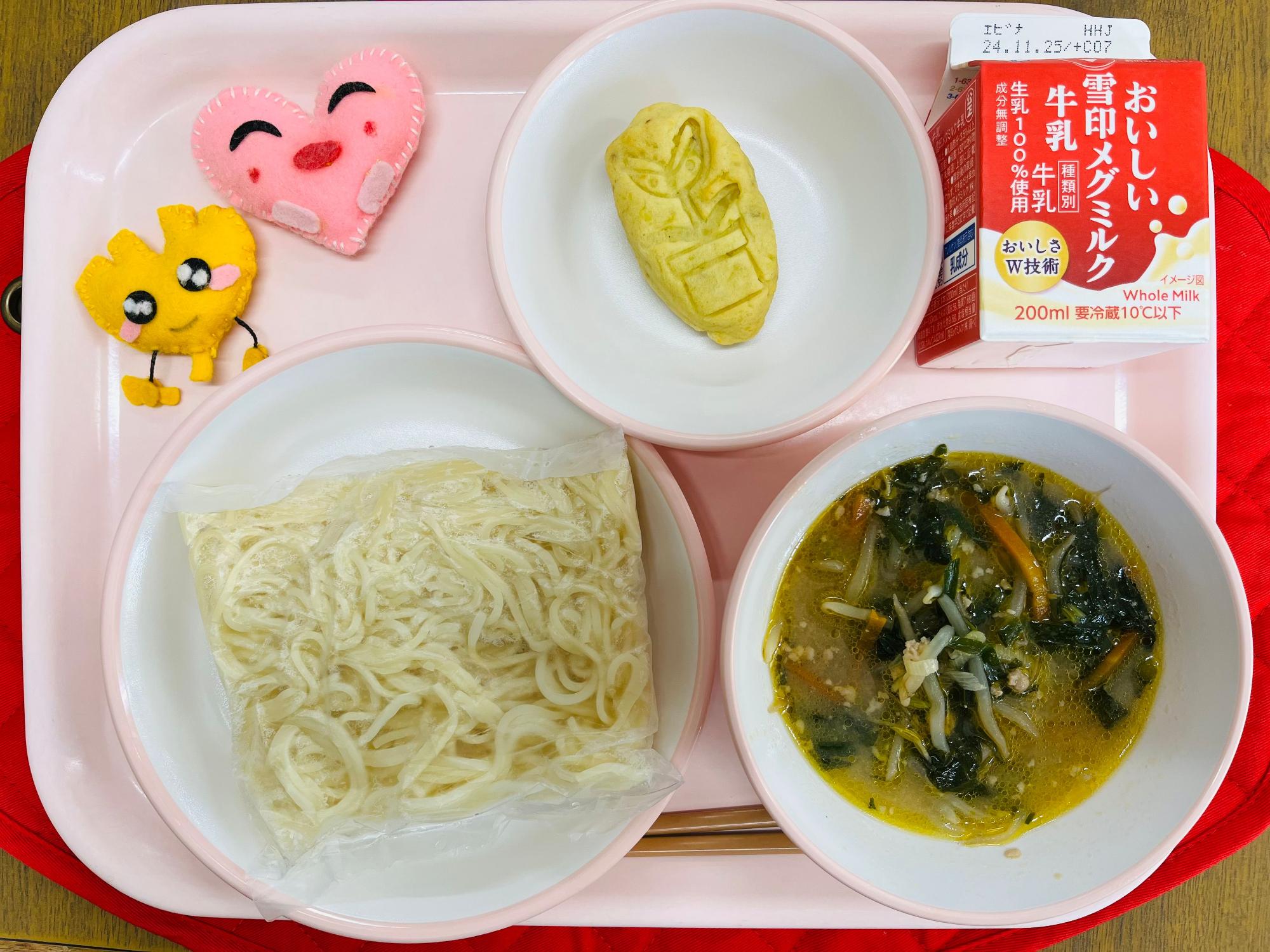 ピンクのトレーに給食が右上から反時計回りで牛乳、スイートポテト、ハートの形をした黄色とピンク色の布製マスコット、麺、みそラーメンのスープと並べられている写真