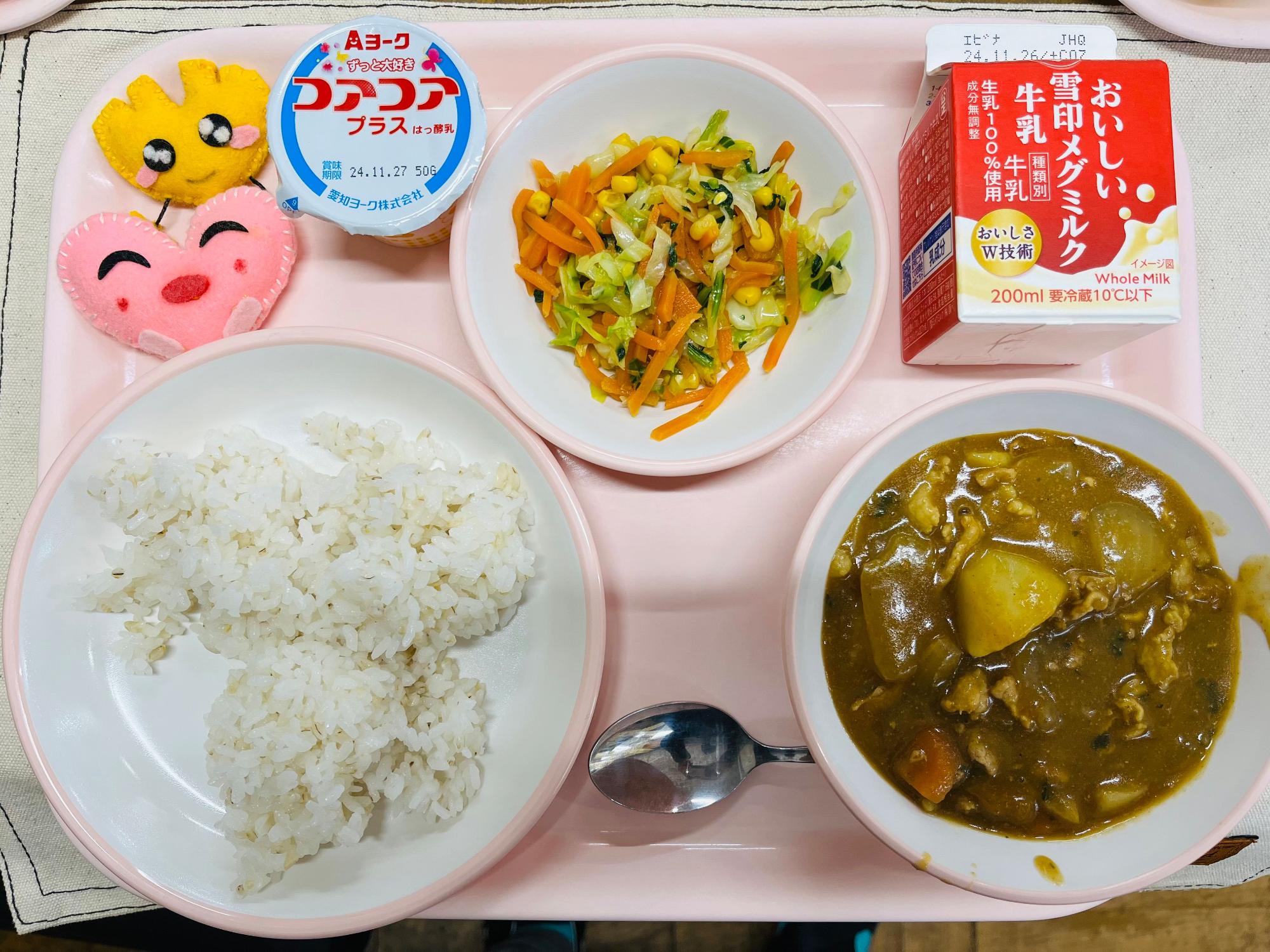 ピンクのトレーに給食が右上から反時計回りで牛乳、コーンソテー、ヨーグルト、ハートの形をした黄色とピンク色の布製マスコット、ごはん、カレーと並べられている写真