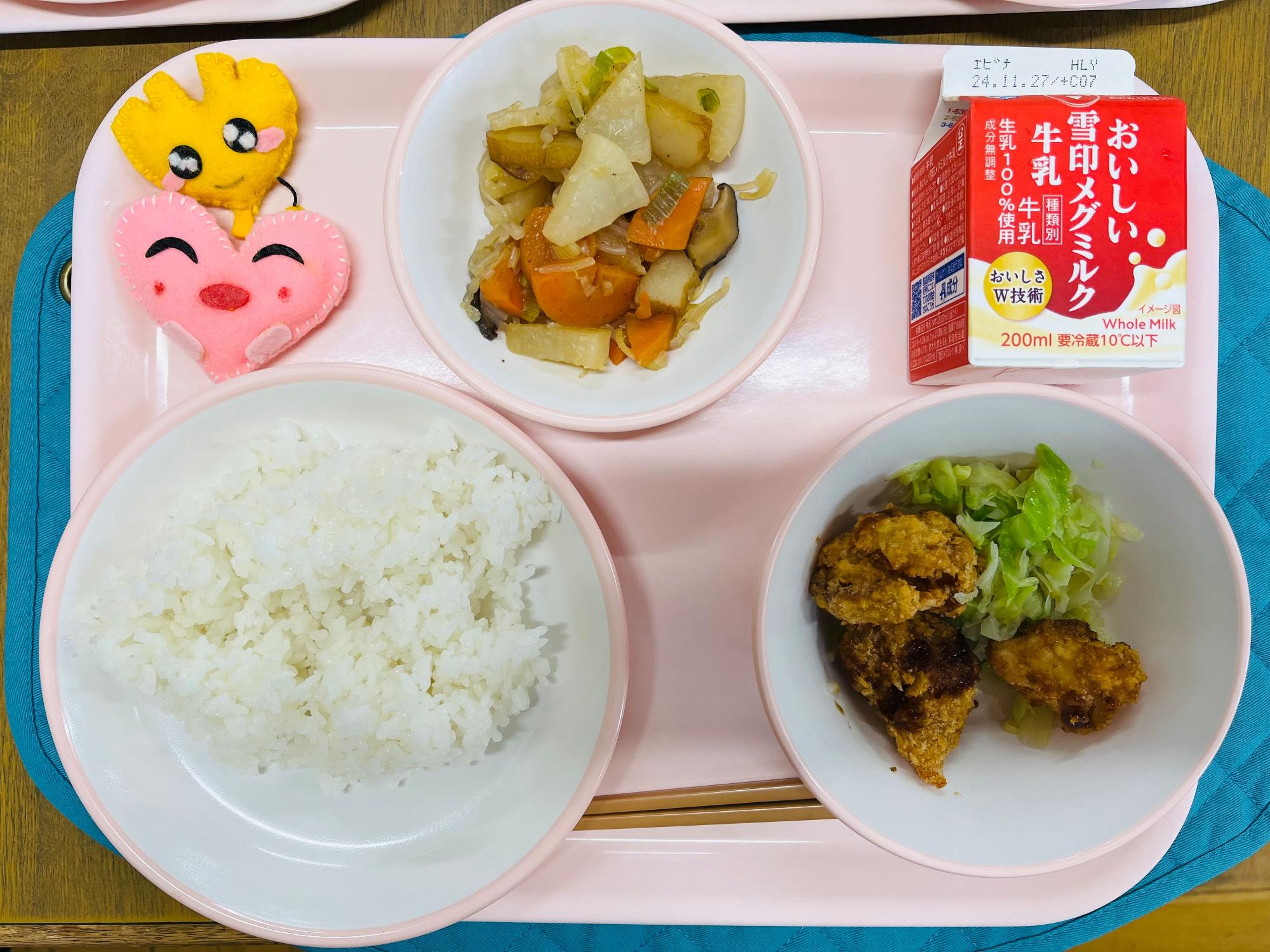ピンクのトレーに給食が右上から反時計回りで牛乳、大根の炒め煮、ハートの形をした黄色とピンク色の布製マスコット、ごはん、キャベツソテーとチキンカツと並べられている写真