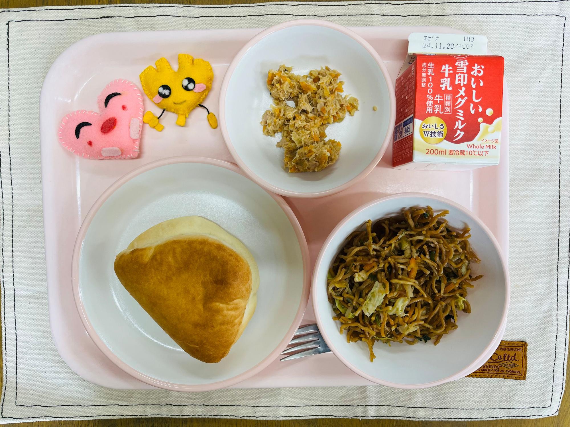 ピンクのトレーに給食が右上から反時計回りで牛乳、ツナサンドの具、ハートの形をした黄色とピンク色の布製マスコット、サンドパン、ソース焼きそばと並べられている写真