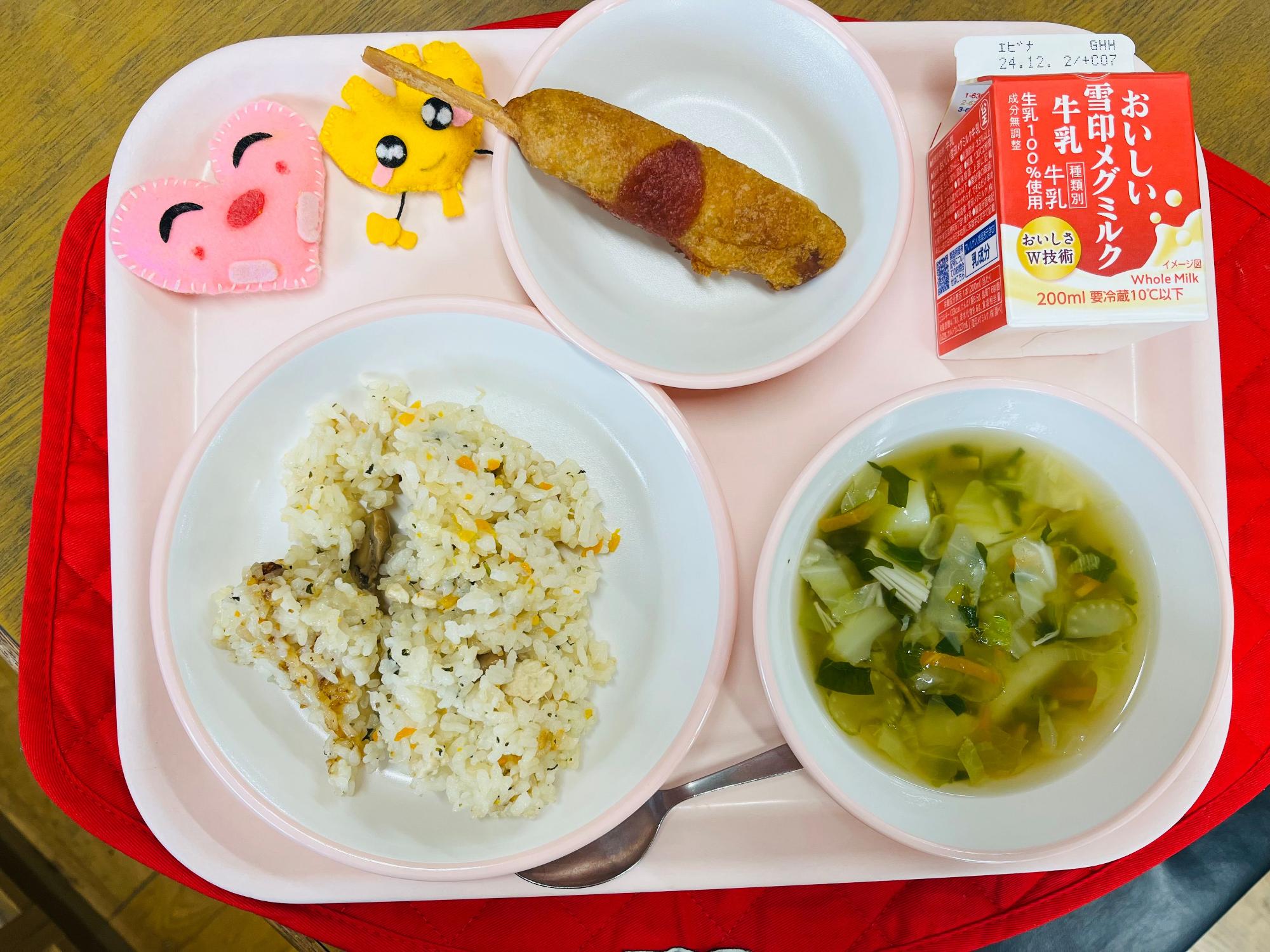 ピンクのトレーに給食が右上から反時計回りで牛乳、アメリカンドック、ハートの形をした黄色とピンク色の布製マスコット、ガーリックピラフ、野菜スープと並べられている写真