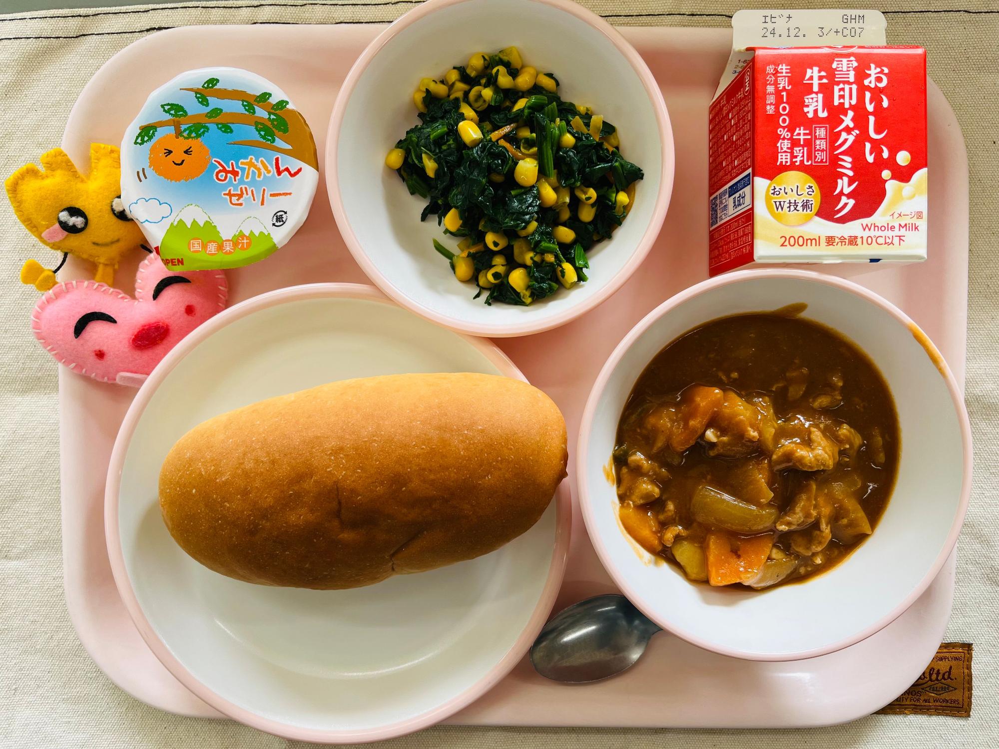 ピンクのトレーに給食が右上から反時計回りで牛乳、ほうれん草のソテー、みかんゼリー、ハートの形をした黄色とピンク色の布製マスコット、コッペパン、ポークシチューと並べられている写真