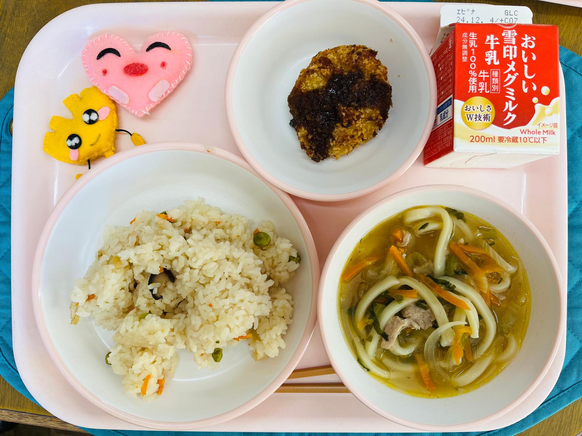 ピンクのトレーに給食が右上から反時計回りで牛乳、カレーコロッケ、ハートの形をした黄色とピンク色の布製マスコット、かやくごはん、にくねぎうどんと並べられている写真