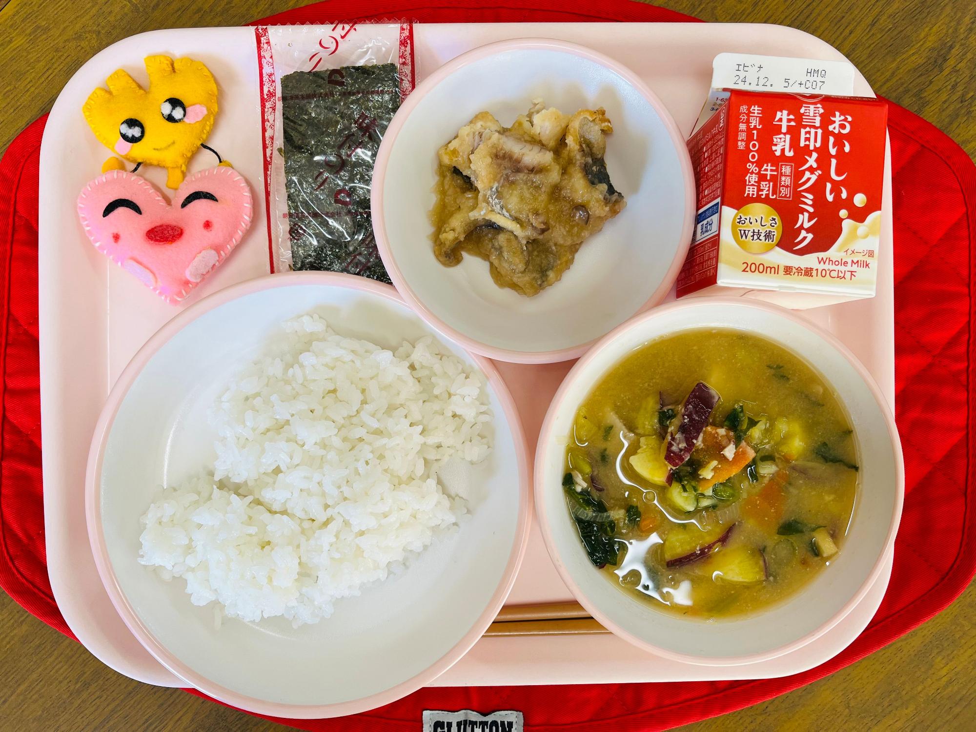ピンクのトレーに給食が右上から反時計回りで牛乳、アジのおろしソースかけ、味付け海苔、ハートの形をした黄色とピンク色の布製マスコット、ごはん、サツマイモの味噌汁と並べられている写真