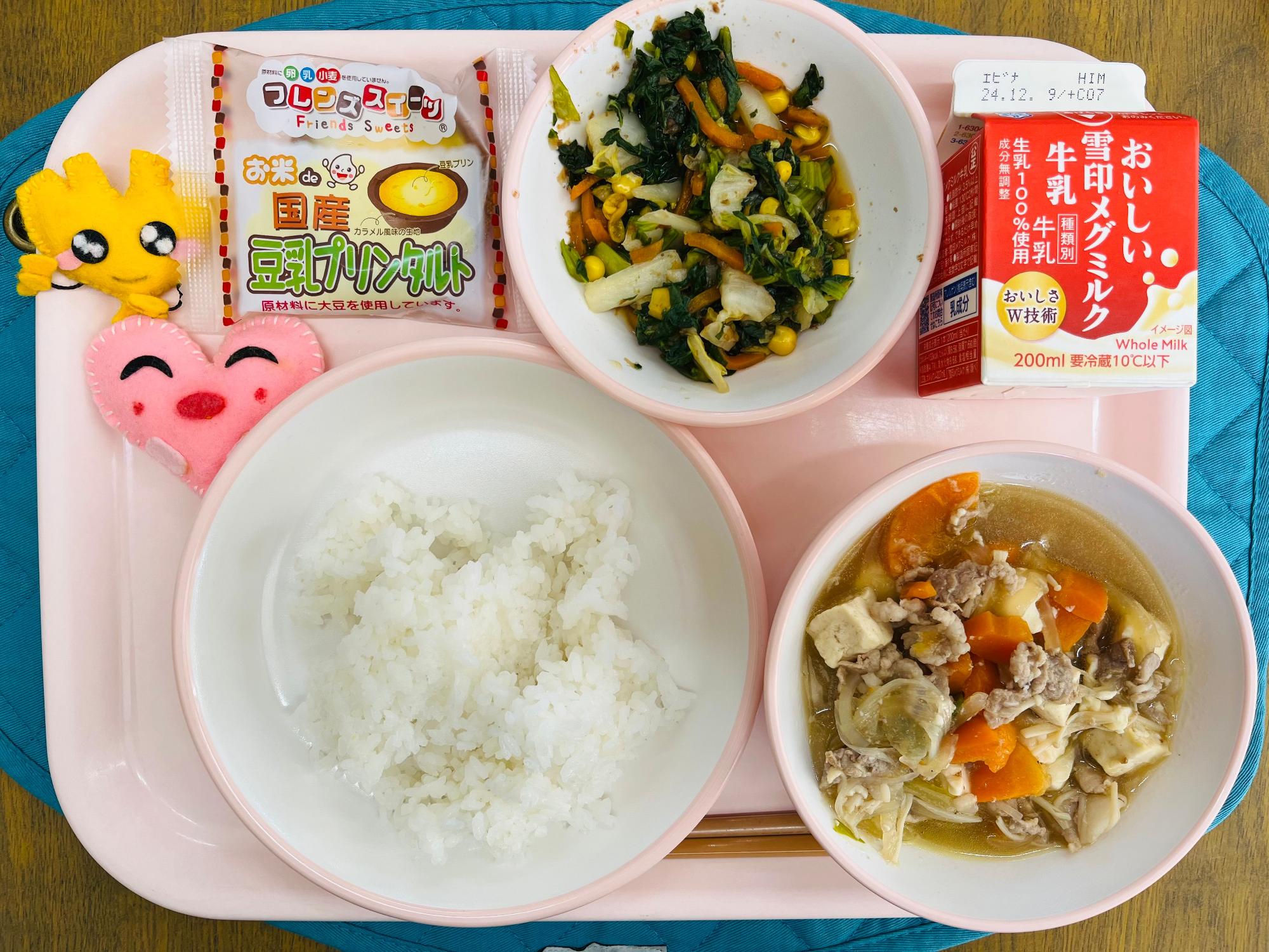 ピンクのトレーに給食が右上から反時計回りで牛乳、カラフルおひたし、豆乳プリン、ハートの形をした黄色とピンク色の布製マスコット、ごはん、肉豆腐と並べられている写真