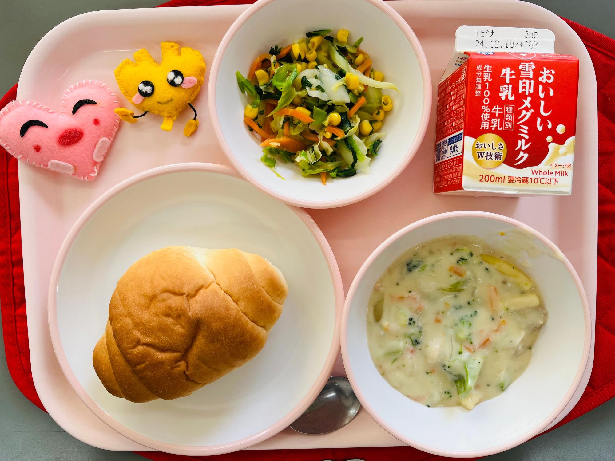 ピンクのトレーに給食が右上から反時計回りで牛乳、野菜ソテー、ハートの形をした黄色とピンク色の布製マスコット、ロールパン、鶏肉とブロッコリーのクリーム煮と並べられている写真